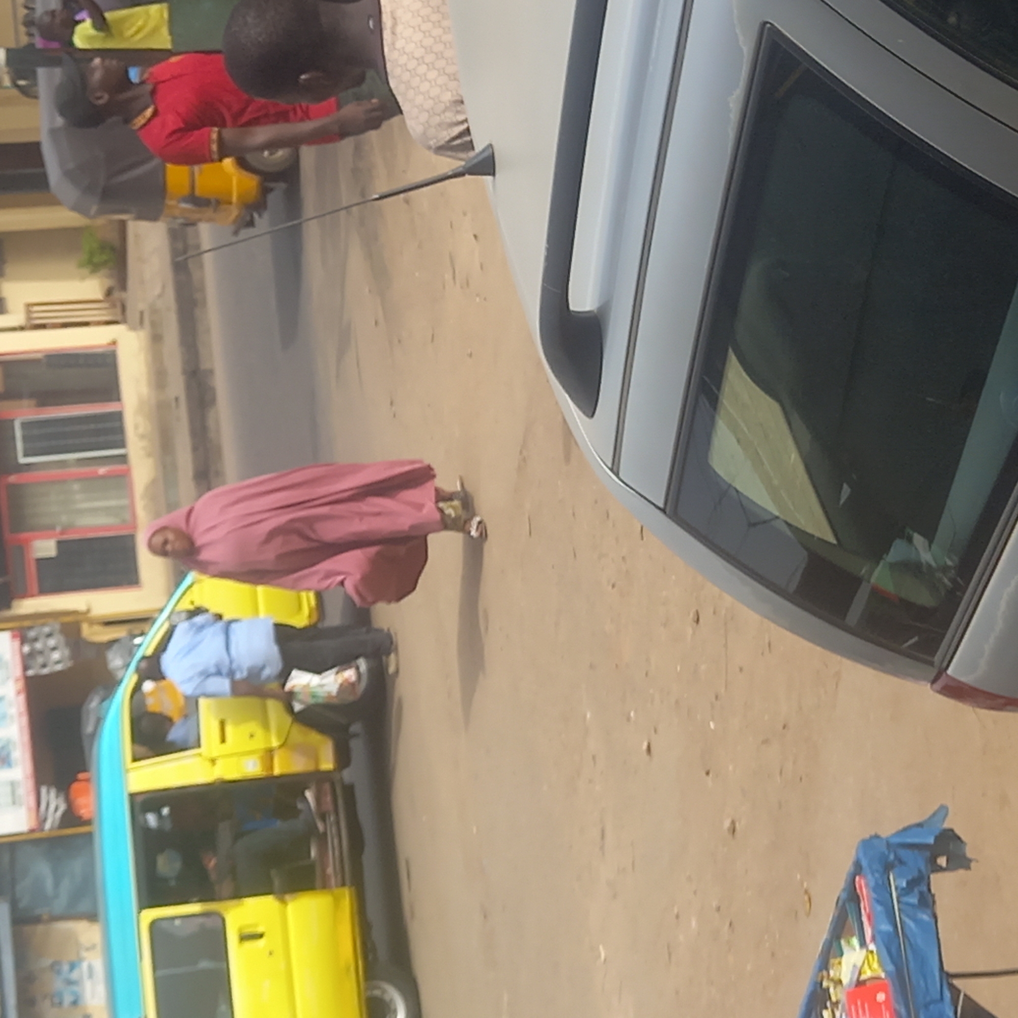 The image depicts a street scene in Dadi Riba, Nigeria. A woman wearing a long, pink garment is walking on the sandy, unpaved road. A yellow and light blue public transport vehicle is nearby, with passengers inside. In the background, there is a building with a red and white facade. A silver car is partially visible on the right side of the frame. On the right foreground, there is a small blue cart containing various items. Other people are visible in the background near a red shirt person.