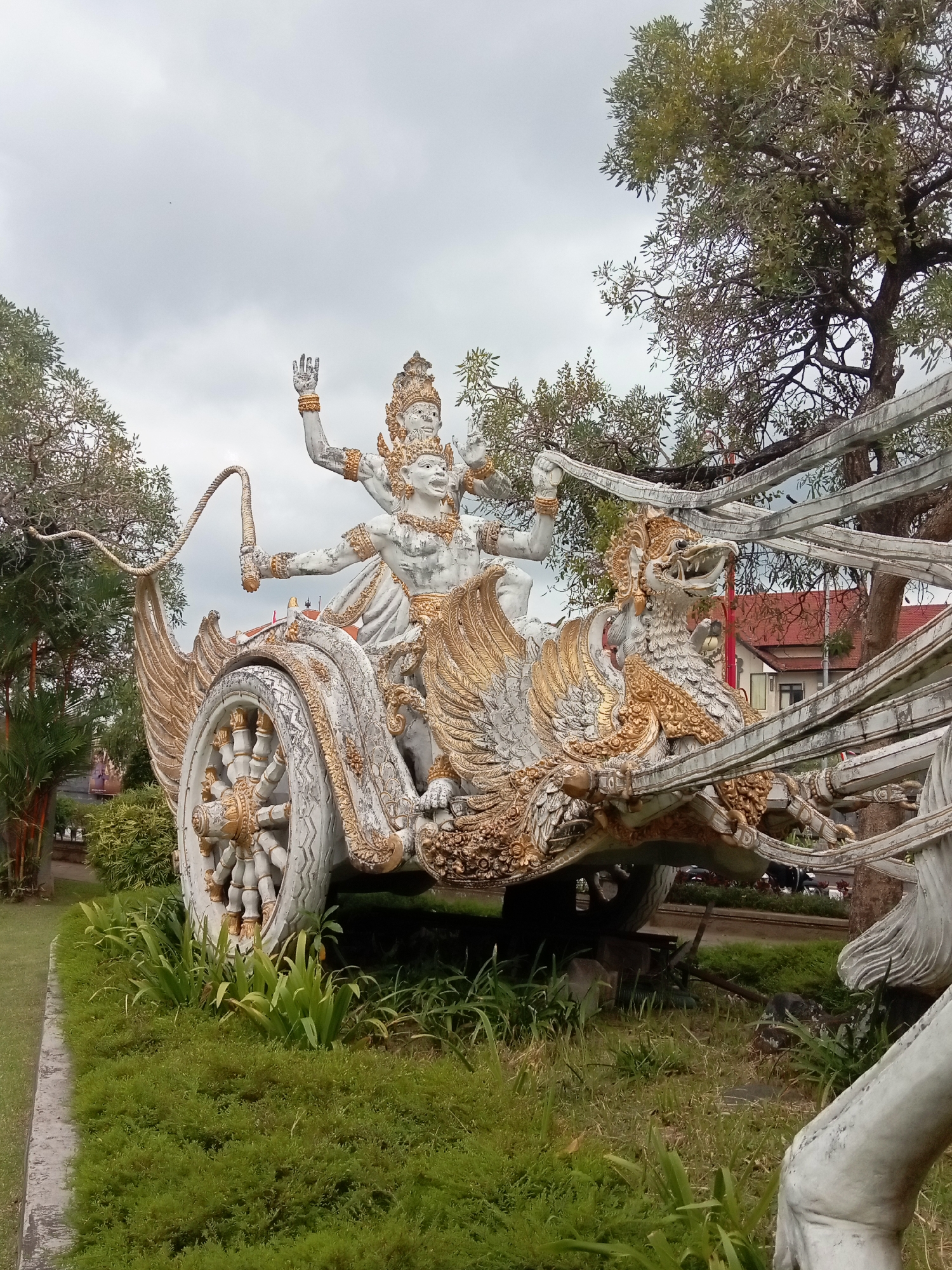 An outdoor, eye-level, medium shot captures a large, ornate statue of a chariot being pulled by winged, mythical creatures. The chariot and the figures are predominantly white with gold accents. The chariot has large, spoked wheels and is adorned with intricate carvings and golden trim. Two multi-armed figures are seated in the chariot, one holding a whip. The creatures pulling the chariot are depicted with open mouths and detailed plumage. The scene is set on a cloudy day, with trees and lush green grass surrounding the statue. In the background, a red-roofed building is partially visible.