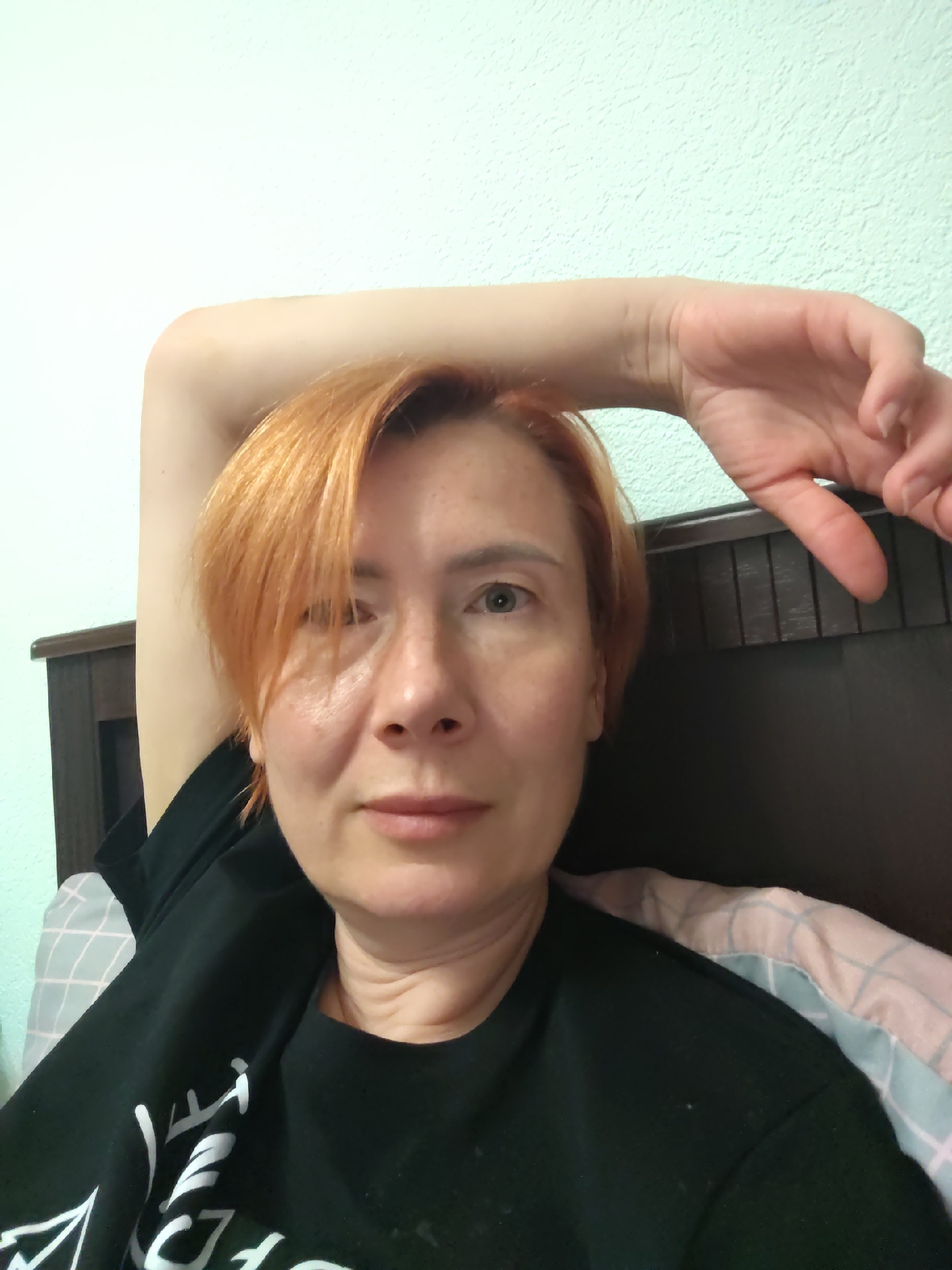 A woman with short, reddish-orange hair is lying in bed, her left arm bent and resting above her head, with her hand extended behind her. She is wearing a black t-shirt with white lettering. She is looking towards the camera with a neutral expression. Her head is positioned against a wooden headboard, and the background consists of a textured light green wall. The bed is covered with a blanket that has a pink and light blue checkered pattern. The image appears to be a selfie taken indoors. Konakovo, Russia is not explicitly visible or discernible in the image itself.