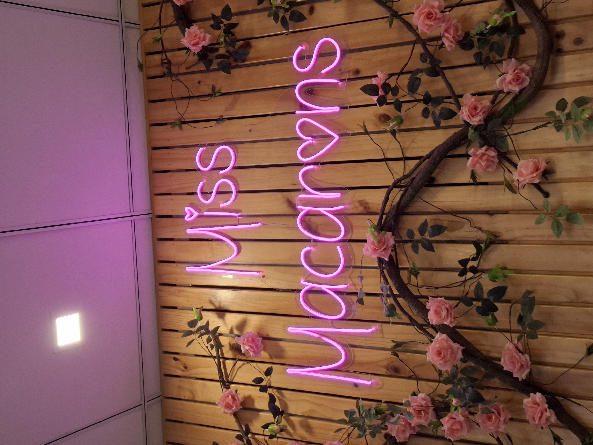 A close-up, eye-level shot captures a neon sign spelling out "Miss Macarons" in bright pink letters, mounted against a wooden plank wall. The sign is adorned with decorative pink roses and twisting vines, which also surround the lower half of the frame. A square, white ceiling light is visible in the upper left corner, casting a cool glow. The overall ambiance suggests a charming, possibly sweet-themed establishment. The location is Unknown, Unknown.
