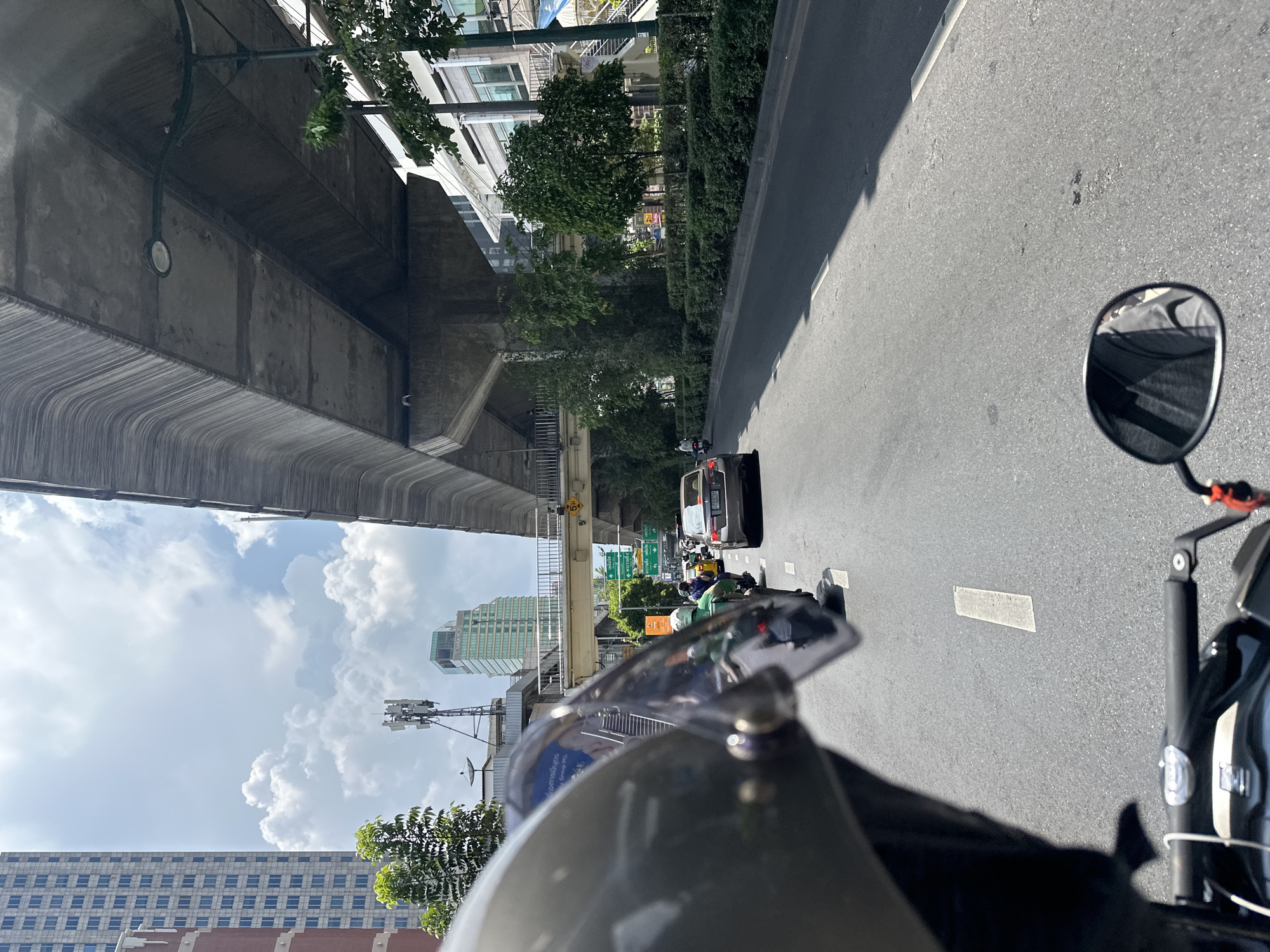 The image captures a street view in Bangkok, Thailand, from the perspective of someone riding a motorcycle. The rider is wearing a helmet with a visible visor. Other vehicles, including a car and other motorcycles, are traveling in the same direction. Buildings and elevated roadways line the street, partially obscured by trees. The sky is visible above, featuring scattered clouds. The motorcycle's rearview mirror is visible in the upper right corner of the frame.