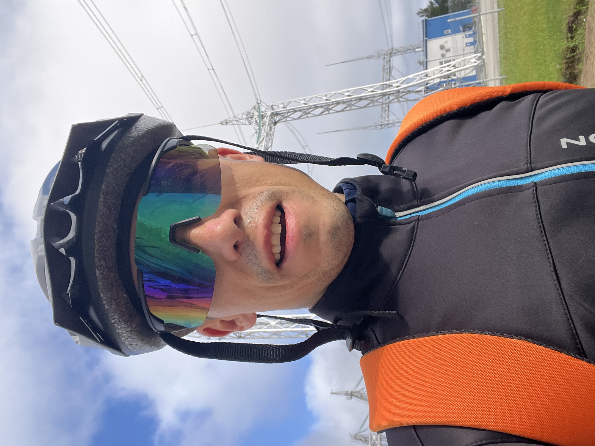 A close-up, selfie-style portrait of a person wearing a bicycle helmet and reflective sunglasses, with an orange backpack strap visible over their shoulder. The person is wearing a black cycling jacket with a blue zipper. In the background, there are tall power transmission towers and a building with blue and white siding. The sky is cloudy with patches of blue. The scene appears to be an outdoor industrial area, likely near a power substation. The location is Chaikovsky, Russia.