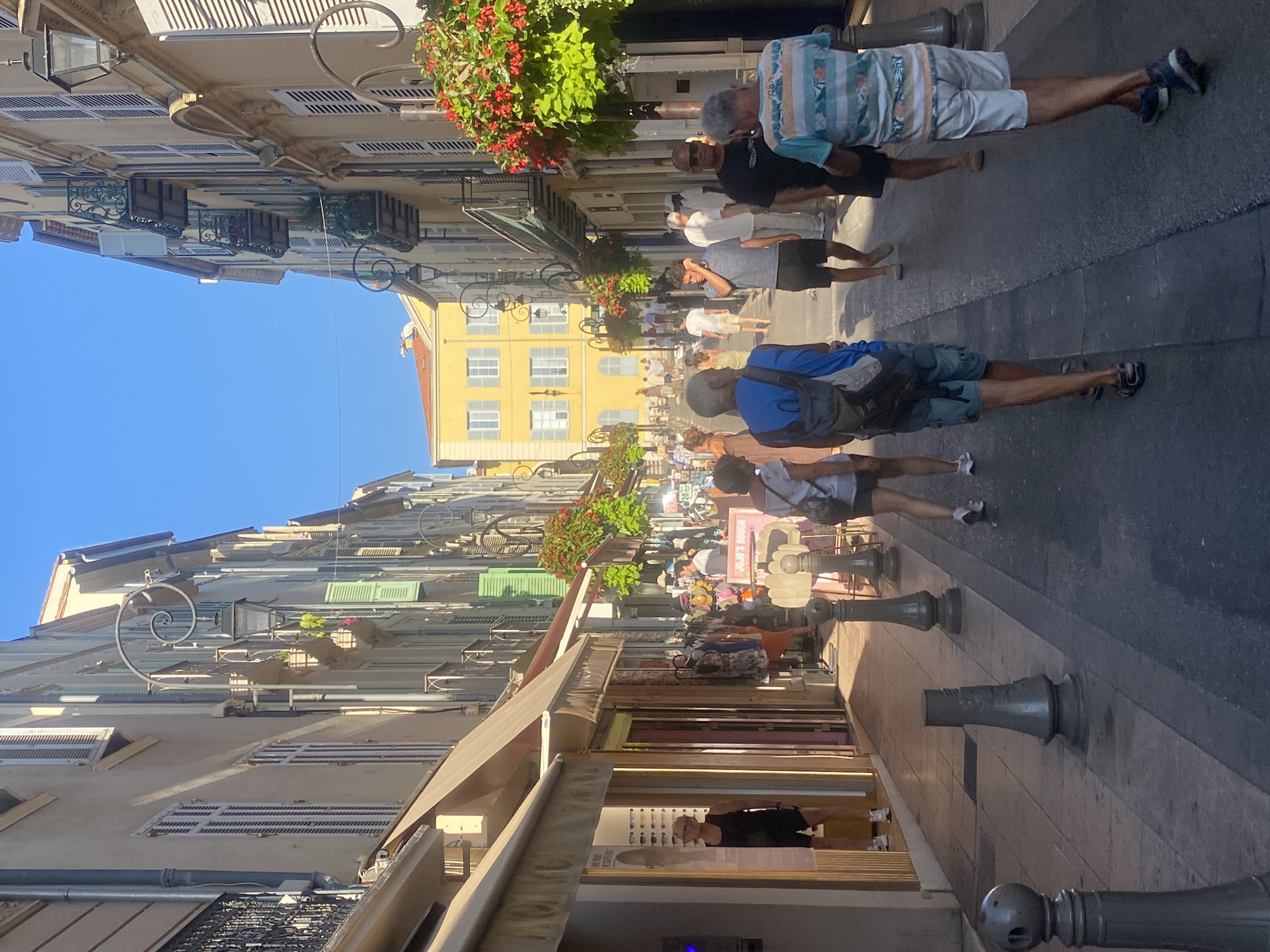 The image depicts a sunlit pedestrian street in Antibes, France, likely during daytime. The street is lined with tall, narrow buildings featuring shuttered windows and balconies, indicative of Mediterranean architecture. Shops with awnings are visible on the left side of the street, some displaying clothing racks and merchandise. On the right, a yellow building stands out in the distance.
Several people are walking along the street. In the foreground, a man wearing a striped t-shirt and white shorts walks away from the camera. To his left, another man in a blue and white striped t-shirt and dark shorts is also walking away. Further down the street, a woman in a white shirt and dark shorts is seen from behind, and a man in a black t-shirt and dark shorts walks towards the camera. Other individuals are scattered throughout the scene, some in small groups, suggesting a leisurely atmosphere.
Decorative elements like hanging flower baskets with red blossoms and ornate streetlights add to the charm of the streetscape. The ground is paved with cobblestones and asphalt. The sky is clear and blue. The overall impression is a vibrant, lively street scene in a European coastal town.