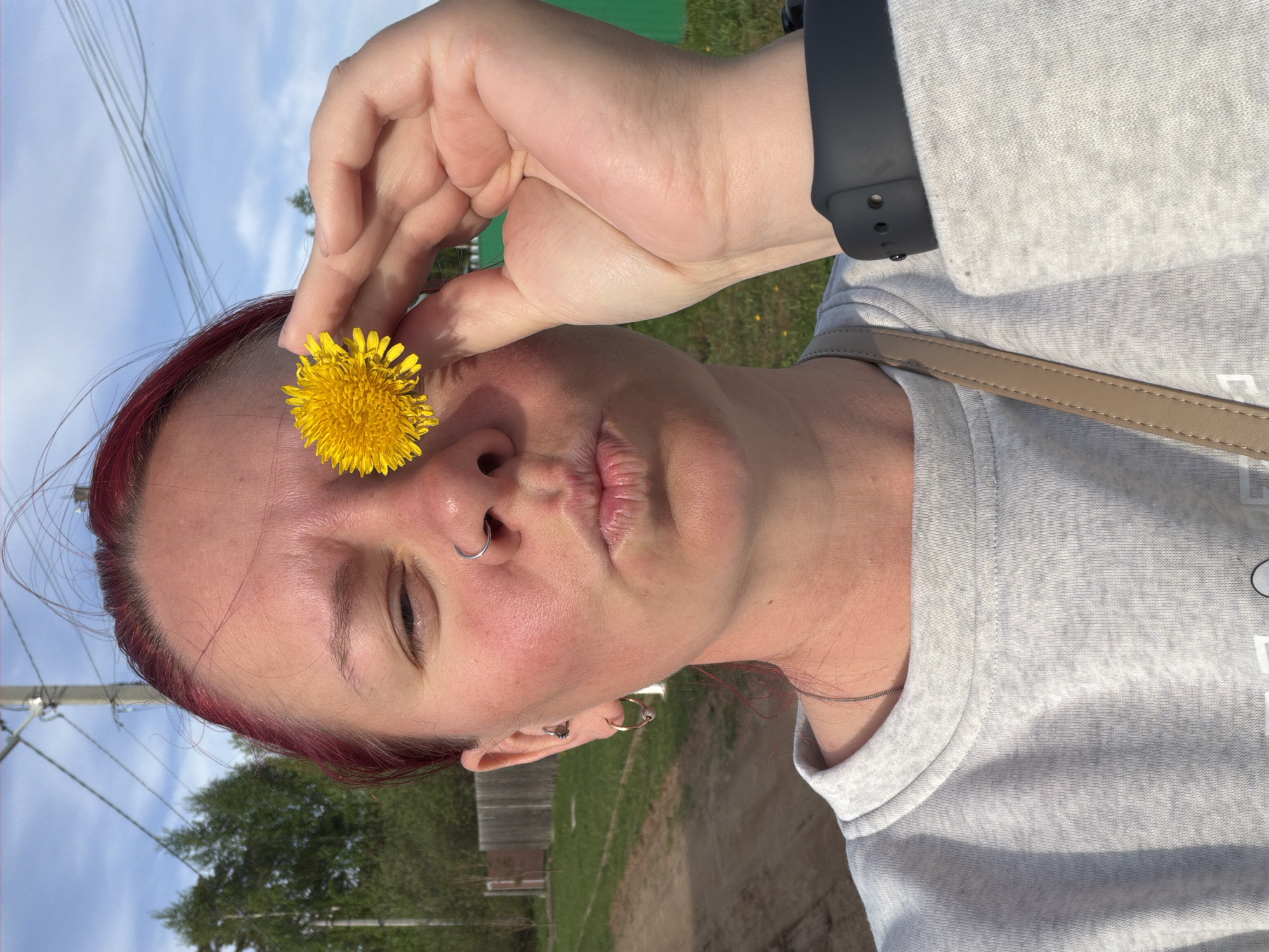 A person is holding a dandelion near their face, outdoors. The person has fair skin, red hair, and is wearing a light grey long-sleeved shirt. A black smart watch is visible on the person's left wrist. The background shows a partially cloudy sky, trees, a road or path, and a wooden fence. The location may be in the city of Chaikovsky, Russia.