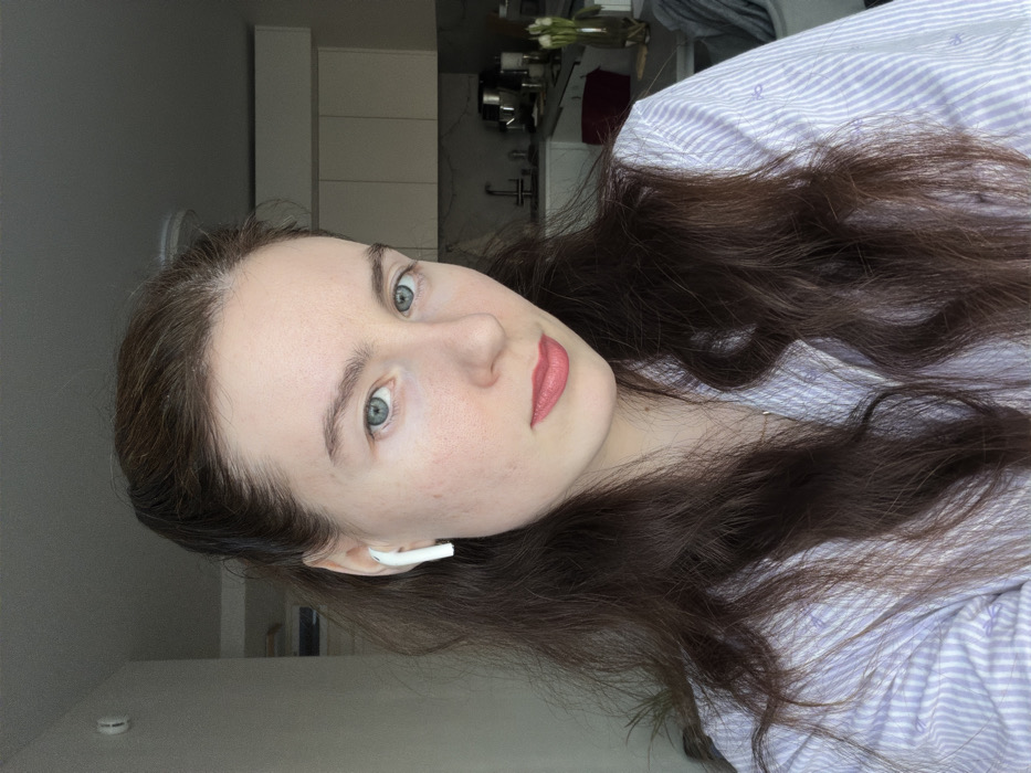 An adult female, with long dark brown hair and blue eyes, is depicted from the upper chest, facing forward with a neutral expression. She wears a light purple and white striped collared shirt and has a white earbud inserted into her left ear. Her lips show a light pink tint. The background is an indoor residential setting, featuring a modern kitchen or kitchenette area. Visible elements include light-colored upper cabinets, a sink with a faucet, and a countertop displaying various kitchen appliances such as a stand mixer and a coffee maker. A vase with white flowers is present on the counter to the right. A grey blanket or throw is partially visible in the lower right background. The scene is located in Ковалёво, Russia.