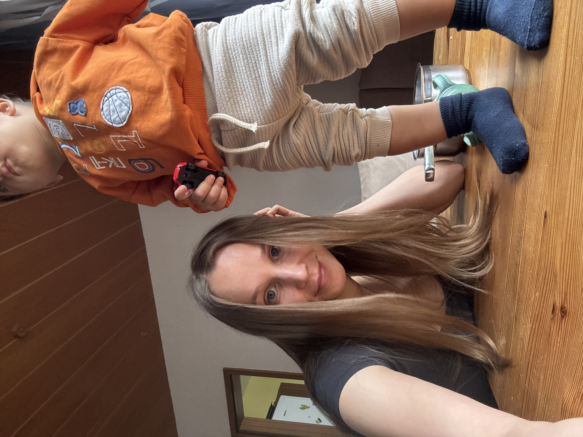 In a domestic indoor setting, a woman with long, light brown hair, wearing a dark t-shirt, is seated at a wooden table. She is looking directly at the camera with a slight smile. To her right, a young child, likely a toddler, is standing on the table, wearing an orange hooded sweatshirt with colorful graphics and light-colored ribbed pants. The child is holding a small red toy car. The child is wearing dark blue socks. In the background, a doorframe leads to another room where a white refrigerator is visible. The overall environment suggests a home in Überherrn, Germany, possibly a kitchen or dining area, with warm-toned wooden ceiling panels and light-colored walls.