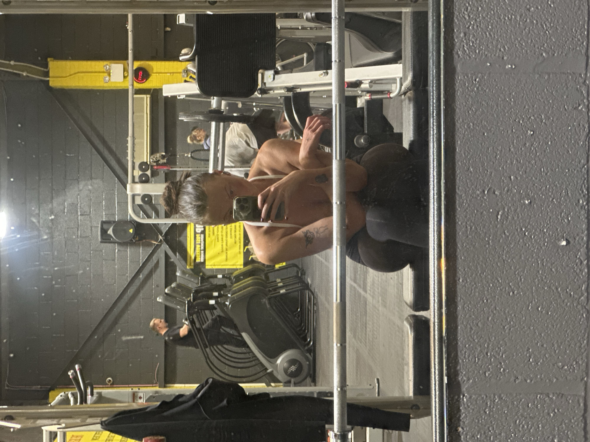 A mirrored selfie in a gym in Tonbridge and Malling, United Kingdom, shows a person holding a phone taking a picture of themselves in the mirror. The person is wearing a white top and black bottoms, with a visible tattoo on their arm. In the background, gym equipment, including weight machines and a rack of chairs, is visible. Another person is present in the background near the weight machines. The walls are gray and black. A yellow structural element with a digital clock is visible in the background, high up on the wall. A black jacket is draped over a surface in the foreground.