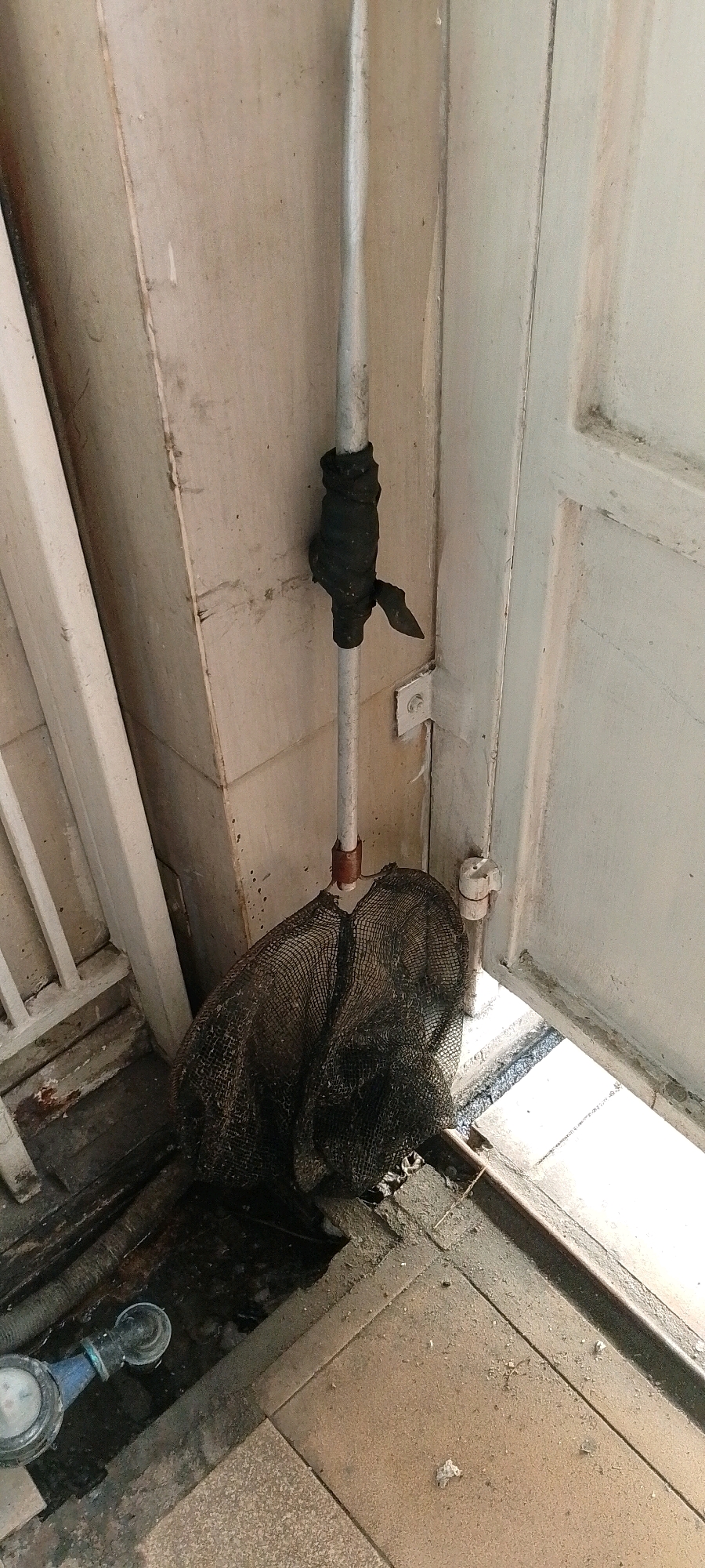 A silver pipe, wrapped with black tape in two places, extends vertically near a white door. At the bottom of the pipe, a dark mesh bag is draped over what appears to be the pipe's connection to a mechanism or support. The background consists of a textured wall and a white door that is ajar, revealing a bright outdoor area. The ground in the foreground is tiled and appears to have some debris. In the lower left corner, a blue and silver valve with a hose is visible. The scene is outdoors, likely in a service or utility area, possibly in the City of Medan, Indonesia.