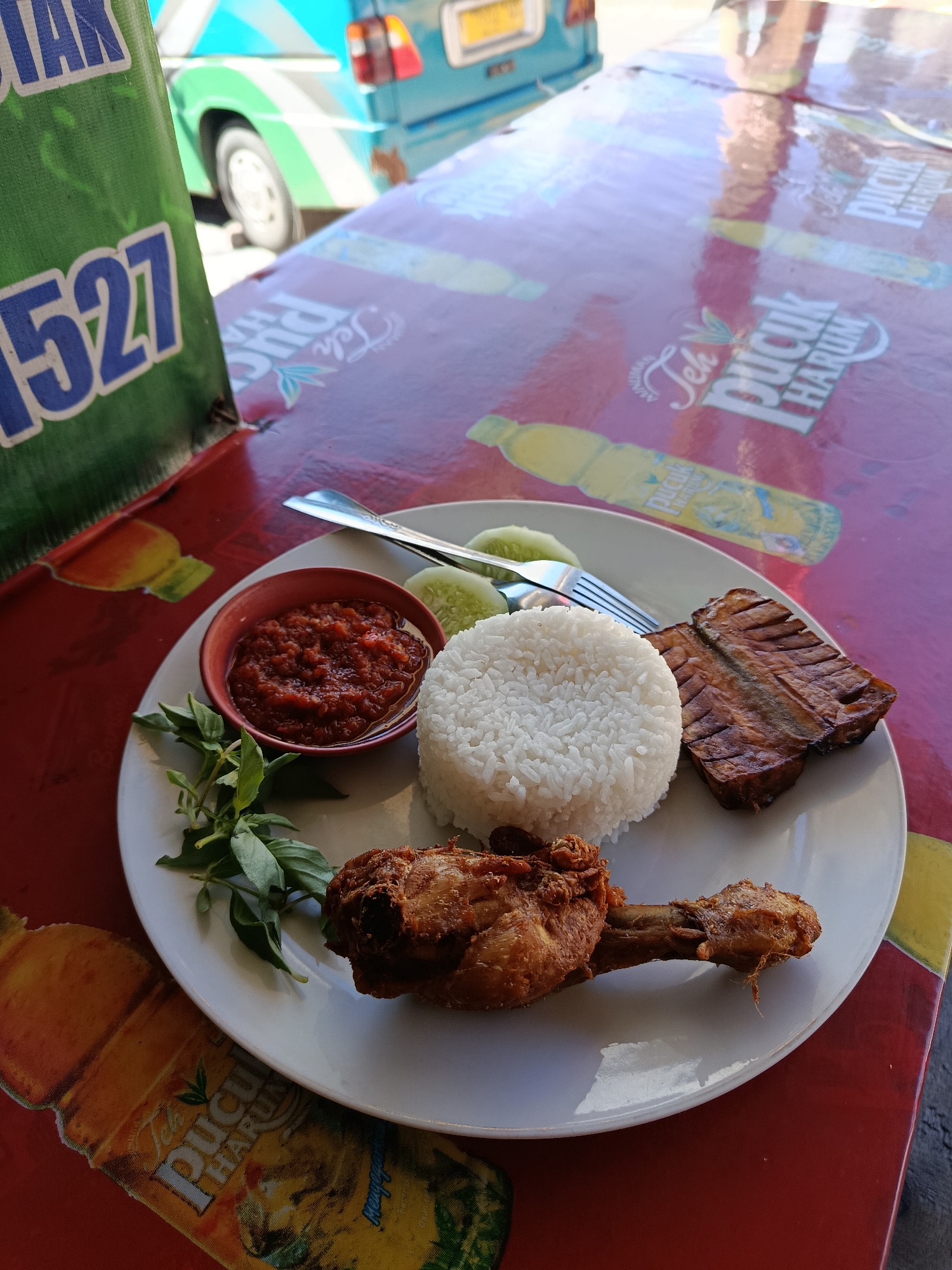 A plate of food sits on a red table with a pattern of beverage advertisements. The plate contains a serving of white rice, a piece of fried chicken, a portion of grilled fish, a small bowl of red chili sauce, and a few slices of cucumber, garnished with fresh green herbs. A fork and spoon rest on the plate. In the background, a blurred green and blue vehicle is visible. The table's surface displays repeated logos for "Teh Pucuk Harum" and a green bag with white lettering, including the number "1527," is placed to the left of the frame. This scene depicts an outdoor meal setting in Cemorolawang, Indonesia.