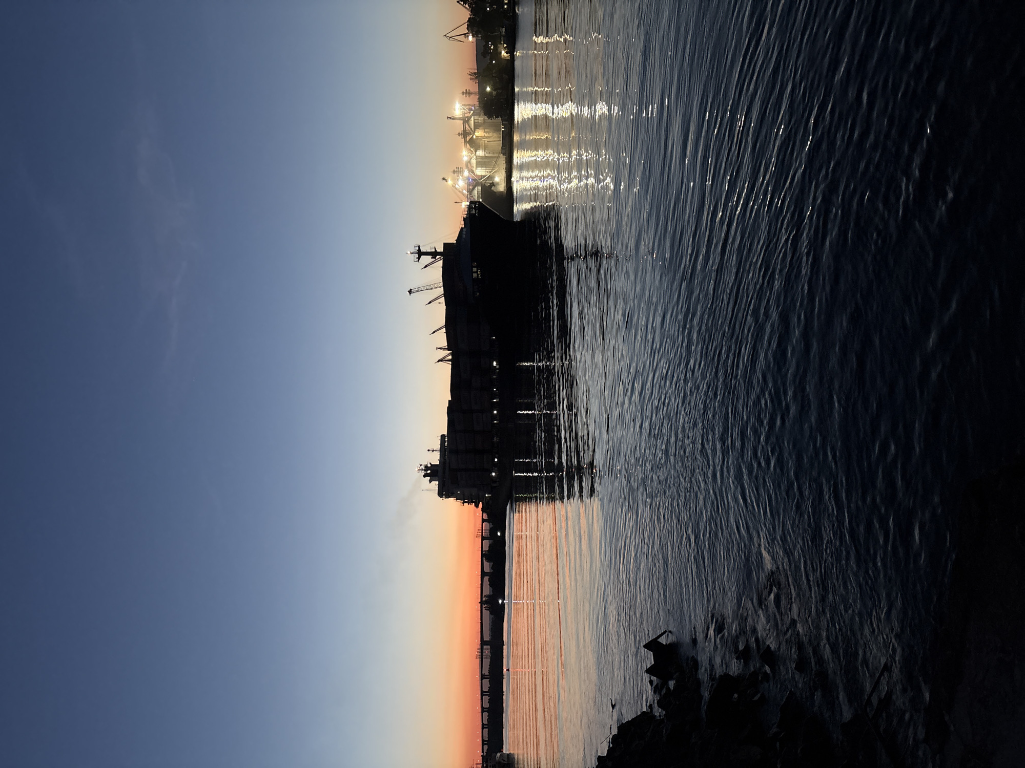 The image depicts a large cargo ship docked in the Port of Varna, Bulgaria at dusk. The vessel is silhouetted against the fading light of the sunset, and its reflection shimmers on the surface of the water. The sky transitions from blue to orange. Lights from the port infrastructure are visible in the background and reflected on the water. The lower right corner of the image shows some dark rocks near the shoreline.