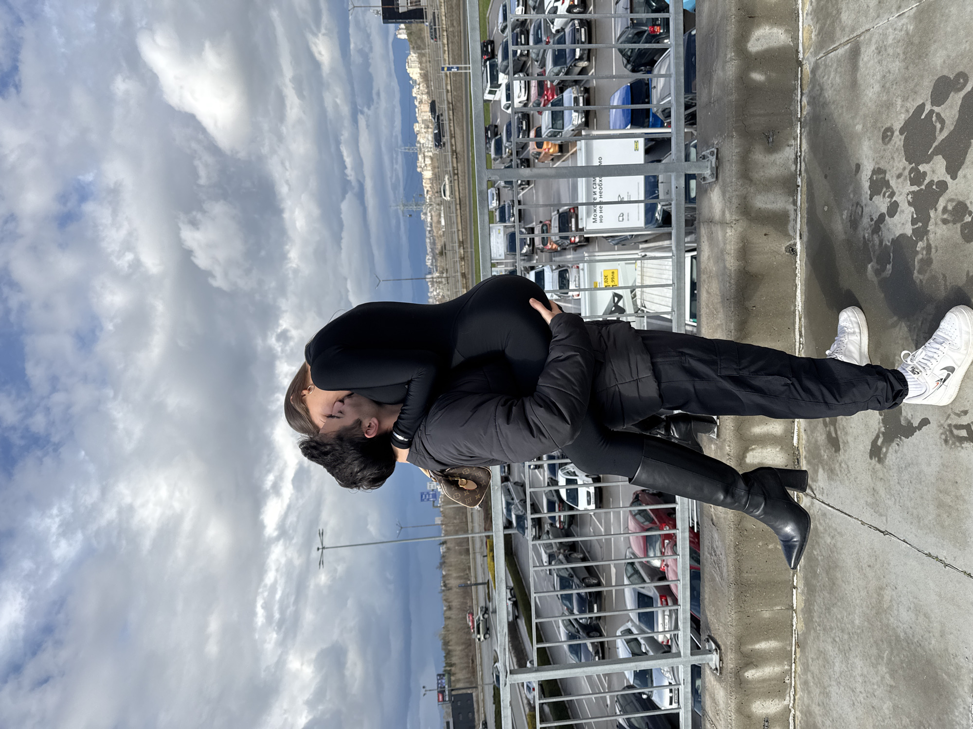 A man and a woman are embracing on the edge of a parking garage rooftop in Sofia, Bulgaria. The woman, wearing a black bodysuit, is leaning her upper body back, supported by the man who is wearing a black jacket and black pants. The woman is also wearing knee-high black boots. The man has dark hair. The woman is wearing sneakers, white with black details. The parking garage overlooks a lot full of parked cars. In the background, there is a city skyline, with a cloudy sky above. The ground around the pair is wet. The woman is carrying a small purse.