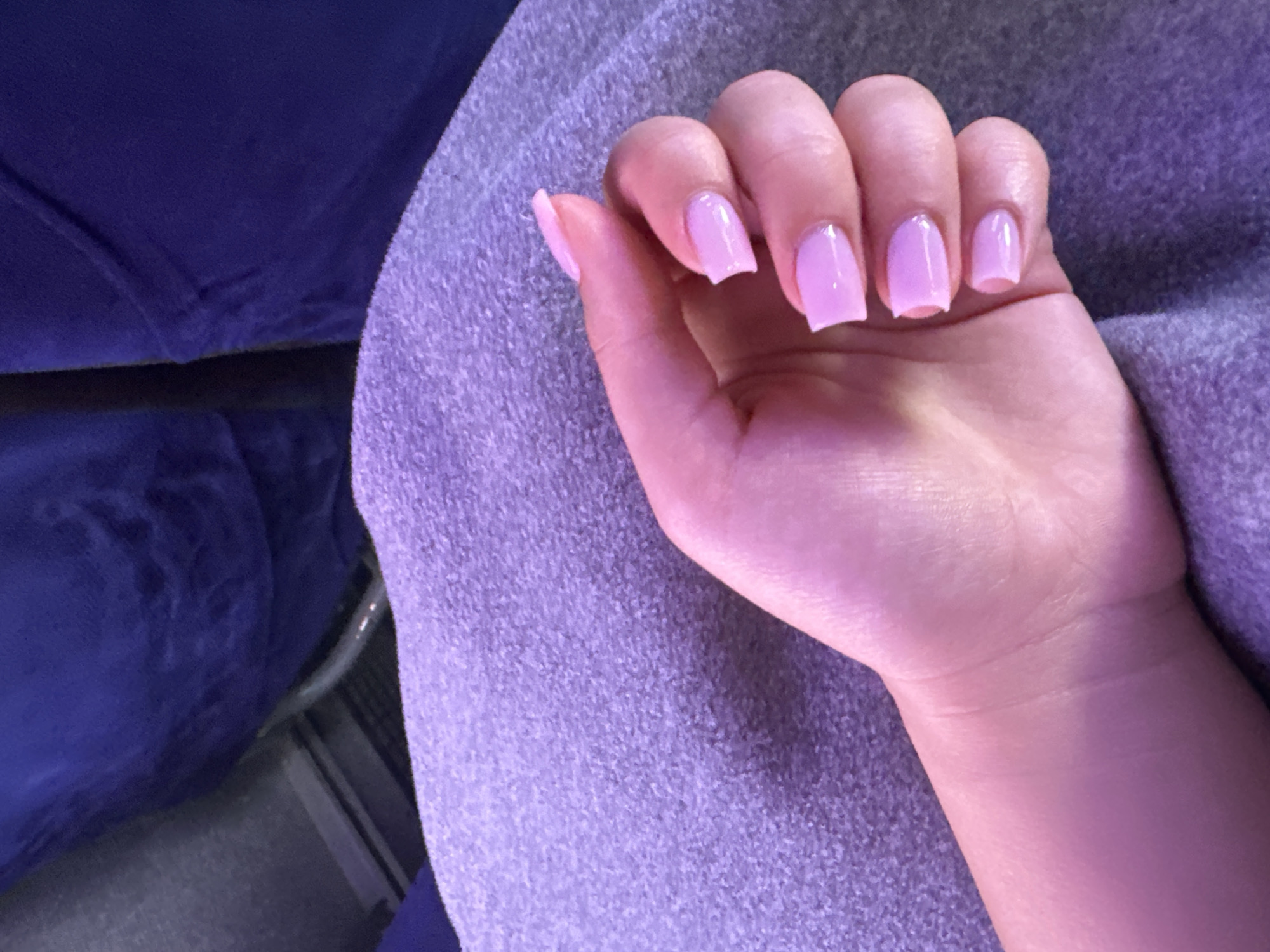 A close-up shot focuses on a person's left hand, adorned with light pink square-shaped fingernails, resting on a textured purple blanket. The hand is positioned to the right of the frame, with fingers gently curled. In the background, dark blue fabric, possibly upholstery from a seat, is visible. The lighting casts a soft, purple hue across the scene. The location is not specified.