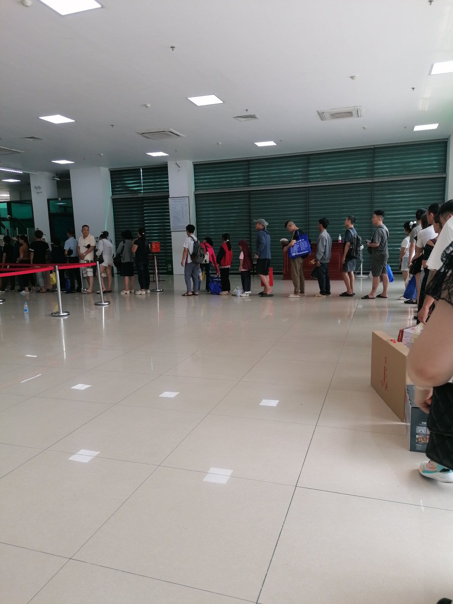 People are standing in a queue inside a large room. The room has a tiled floor and a white ceiling with square lights. The queue is formed using red stanchions and ropes. People are of varying ages and are dressed casually. Some are carrying bags or backpacks. There is a desk at the end of the queue. The location is likely a government office or service center in Lao Cai Ward, Vietnam.