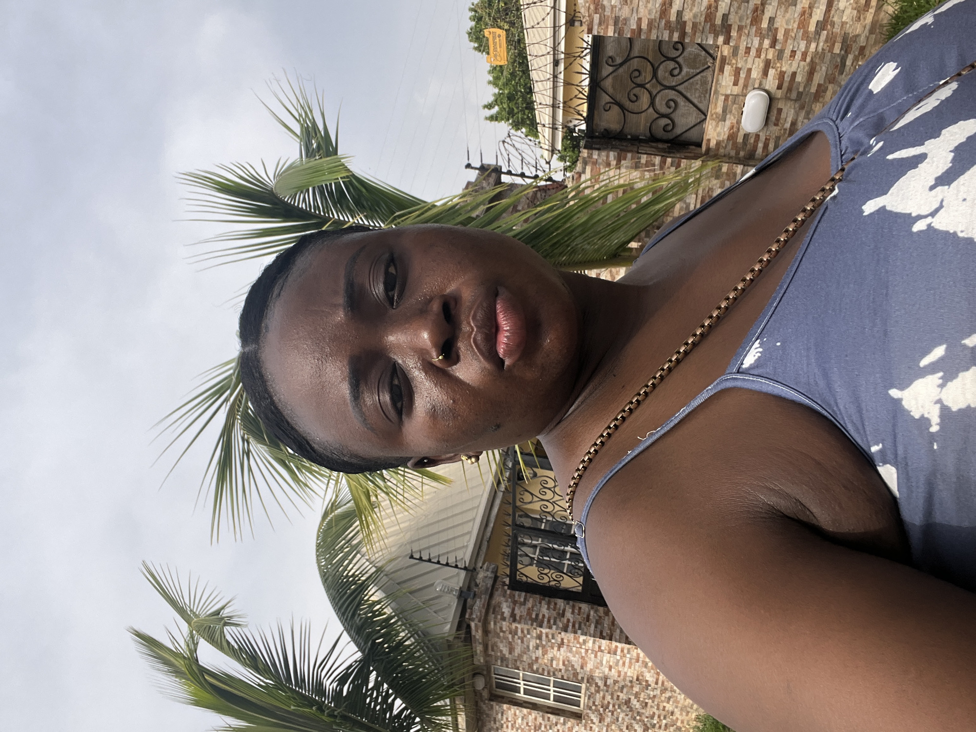 A medium shot captures a Black woman with short hair, a nose ring, and a gold chain, positioned at an angle that includes palm trees and a building in the background. The building features a window with a decorative wrought iron grill, a stone facade, and a wall-mounted light fixture. The sky is overcast. The location is possibly in Abuja, Nigeria.