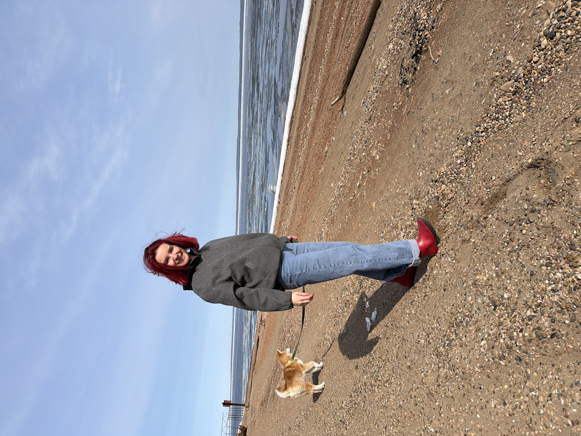 A woman with red hair is walking a Shiba Inu dog on a leash on a sandy, pebbly beach. She is wearing a gray jacket, blue jeans, and red boots. The dog is light orange and white. In the background, there is a body of water with some ice floating on it, and a blue sky with wispy clouds. The location is Chaikovsky, Russia.
