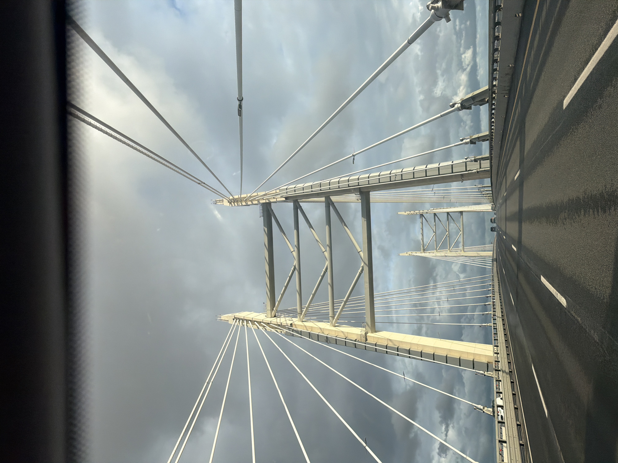 The image shows a view from a vehicle traveling on a cable-stayed bridge. The perspective is looking upwards and diagonally, with the road surface of the bridge dominating the right side of the frame. The road is asphalt with white lane markings. On the left and center of the image, the intricate structure of the bridge is visible, featuring tall white pylons and a network of suspension cables. These cables fan out from the pylons to support the bridge deck. The sky above is filled with dramatic, dark grey clouds, suggesting overcast or stormy weather. The lighting indicates it is likely daytime, though the cloud cover creates a subdued atmosphere. The image has a somewhat abstract quality due to the close-up view of the bridge's engineering and the dramatic sky. No people are visible. The scene evokes a sense of scale and the power of engineering against a turbulent natural backdrop.