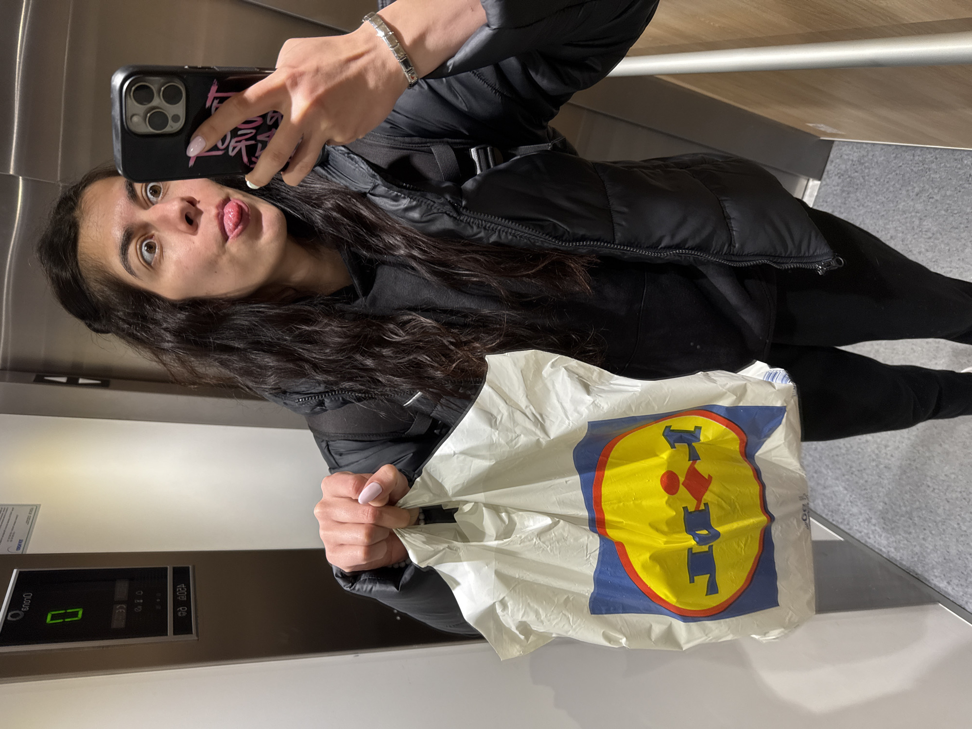 A woman is taking a selfie in what appears to be an elevator. She is holding a Lidl grocery bag, suggesting she may have just been shopping. The elevator's control panel indicates floor "0". Given the presence of a Lidl bag, the location is likely somewhere in the city of Sofia, Bulgaria, where Lidl stores are prevalent. The woman is wearing a black jacket and black pants. Her phone case is black with pink writing. She is sticking her tongue out.