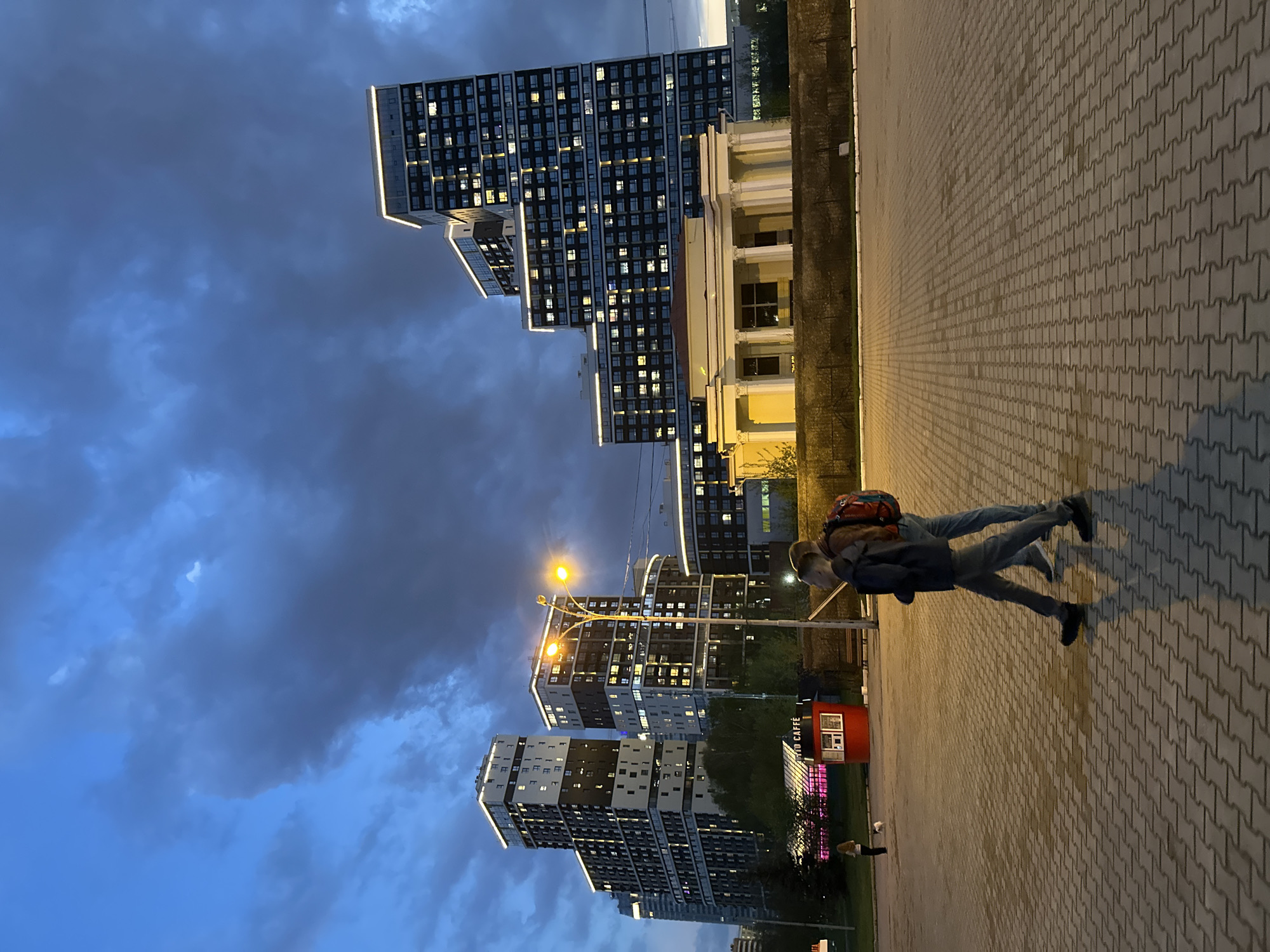 Two people stand on a paved walkway next to modern buildings in Yekaterinburg, Russia. The sky is a mix of blue and dark clouds. The buildings are tall, with many lit windows. Streetlights illuminate the scene. A small red booth labeled "Tutto Caffe" stands nearby.