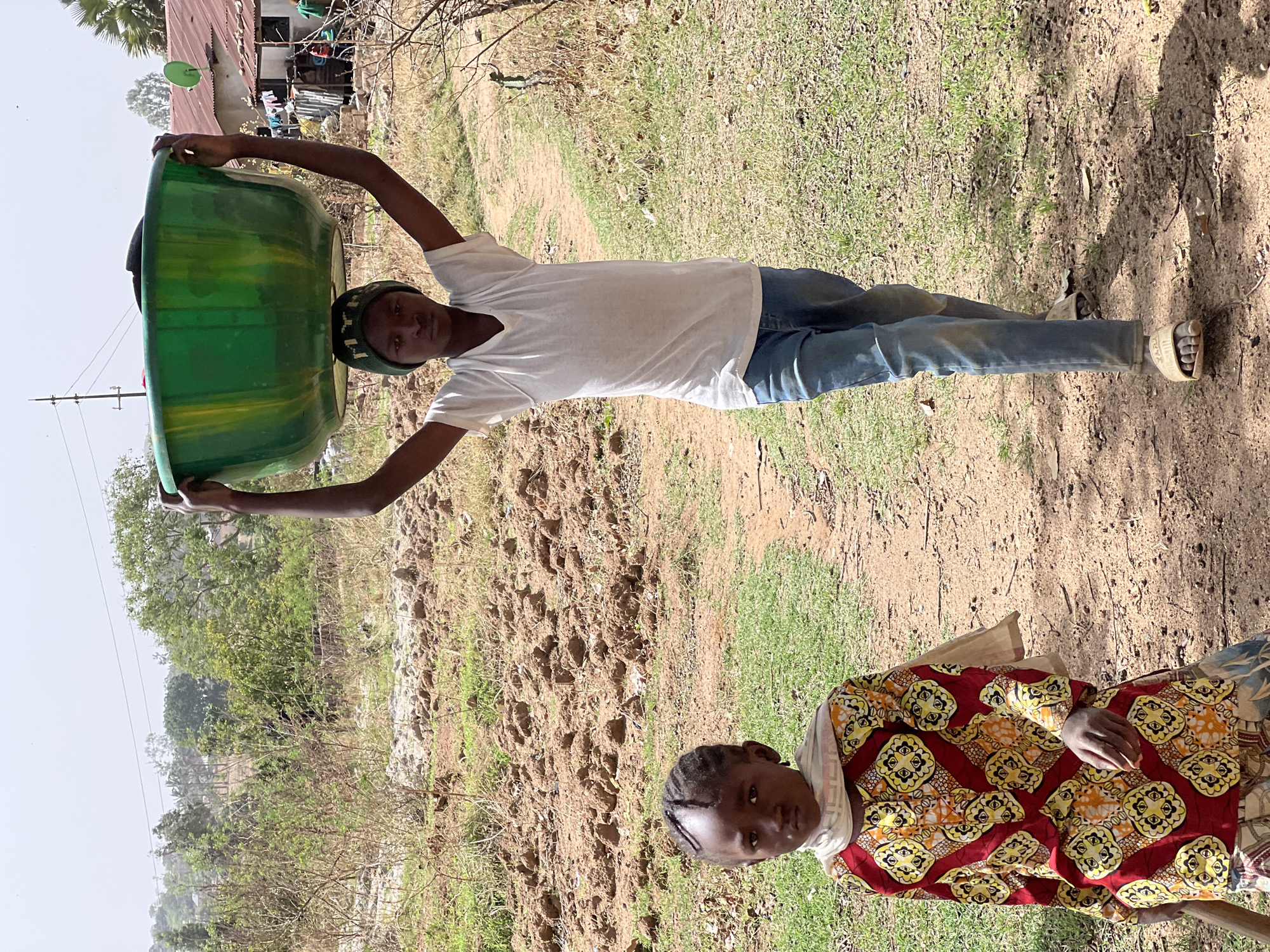 A medium-shot depicts two individuals outside in a rural or semi-rural area, likely in Bukuru, Nigeria, given the prompt's location specification. A young man is seen holding a large, green plastic tub overhead, his arms outstretched. He is wearing a white t-shirt, blue jeans, a camouflage-patterned cap, and sandals. To the bottom right, a younger girl is seen in a colorful patterned dress. She is looking directly at the camera. The environment includes a dirt path, patches of grass, and sparse vegetation. In the background, a building with a red roof is visible. The sky is overcast.