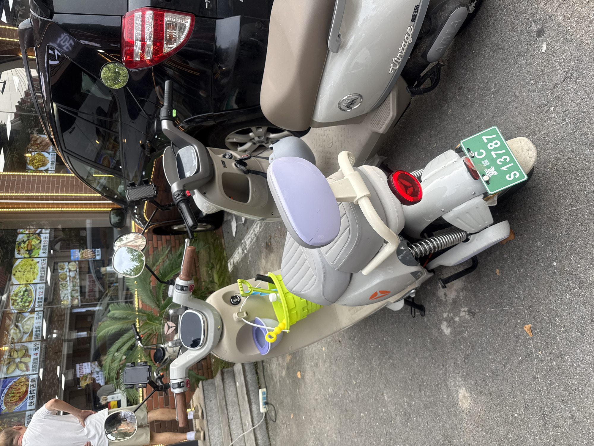 The image shows a street scene in the city of Qinhuangdao, China. In the foreground, two electric scooters are parked on asphalt pavement. The scooter on the left is a light beige and grey model with a green basket attached to the front, containing children's toys and a bottle of water. The scooter on the right is a lighter color with a brown seat and "Vintage" written on its side. Behind the scooters, a black SUV is parked. In the background, a restaurant with a large glass window displays various food items. A man in a white t-shirt and khaki shorts is visible on the left side of the image, standing on steps leading up to the restaurant. The environment appears to be a commercial area.