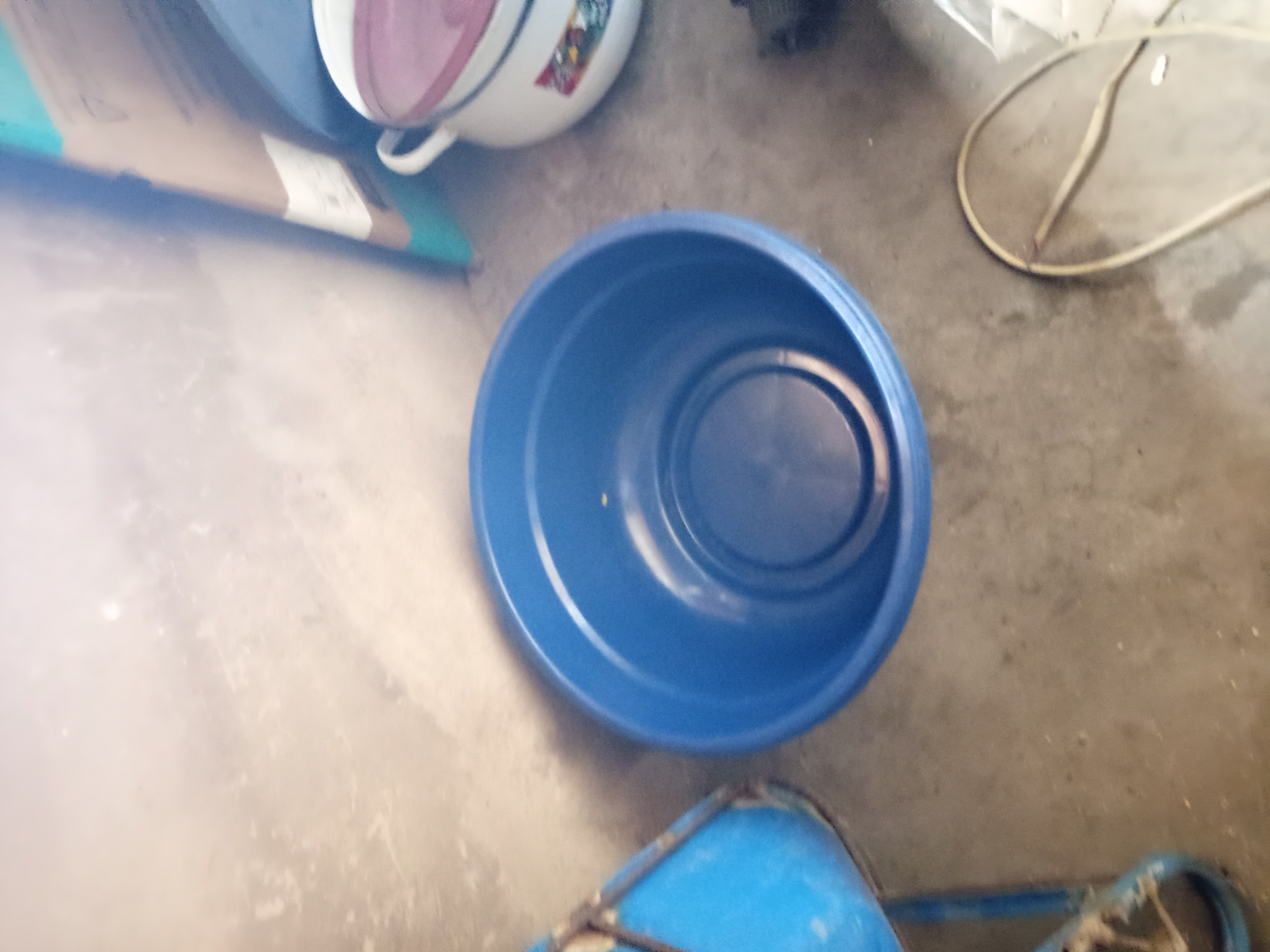 In Jauro Yinu, Nigeria, a medium shot captures a floor scene. In the foreground, a large, bright blue plastic basin is centrally placed. The basin has visible concentric rings inside and a slightly elevated inner base. To the left of the blue basin, a portion of a blue bucket on a metal stand is visible. To the right of the blue basin, a white pot with a pink lid and decorative floral patterns is partially in frame. The pot has two white handles. In the background, a light-colored, textured wall is present, with a vertical brown cardboard box leaning against it. A coiled electrical cord lies on the concrete floor in the lower right corner of the image. The overall lighting suggests an indoor setting, possibly a storage area or a workshop.