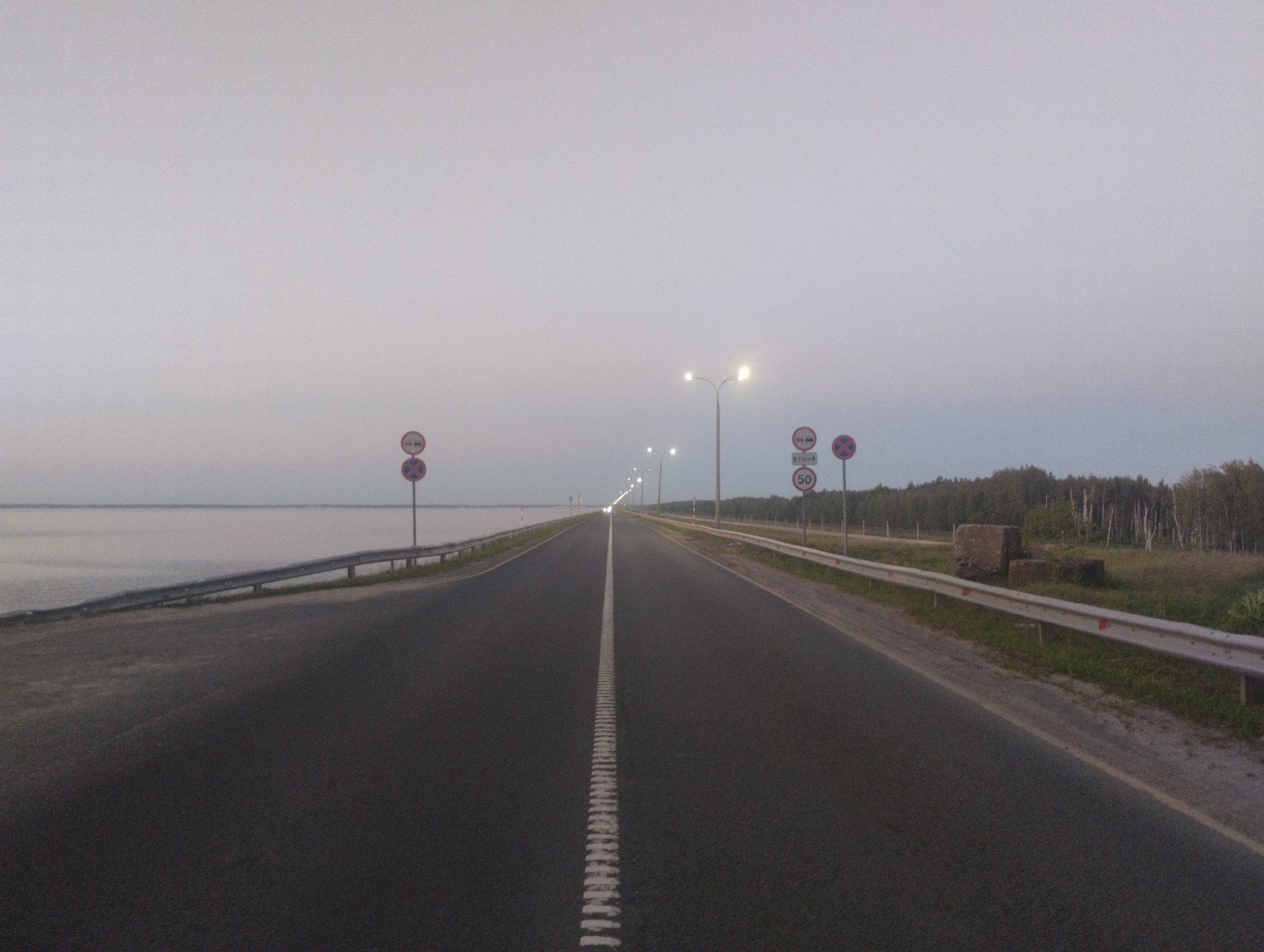 The image shows a straight, paved road in the city of Zavolzhye, Russia, likely at dusk or dawn due to the ambient light. The road has a white dashed line down the center and extends into the distance where it is illuminated by streetlights. On the left side of the road, there is a body of water, possibly a river or lake, with a metal guardrail along the edge. On the right side of the road, there is a grassy area with trees in the background and more guardrails. Several traffic signs are visible: a "no entry" sign and a speed limit sign indicating 50 km/h on the right side, and two "no entry" signs on the left. There are no people or vehicles visible in the image. The sky is overcast and light grey.