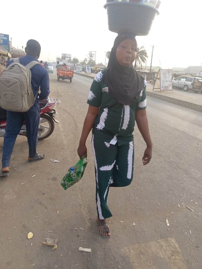 This image depicts a vibrant street scene in Alizaga, Nigeria, under what appears to be bright, hazy natural light, possibly indicating late afternoon or early morning.
In the foreground, a woman walks towards the right side of the frame, balancing a large, dark basin or bowl on her head, characteristic of street vendors. She is dressed in a dark green two-piece outfit featuring white abstract brushstroke patterns and wears a black headscarf. In her left hand, she carries a green bag containing a plastic bottle. Her mouth is slightly open, suggesting she might be speaking or calling out to potential customers. She is wearing simple sandals.
To the left and slightly behind her, a man stands with his back mostly to the camera. He wears a blue long-sleeved shirt, blue trousers, and sandals. A light brown backpack with a prominent "Champion" logo is visible on his back. He is positioned next to a red motorcycle or scooter, only partially visible.
The setting is an asphalt road with some litter scattered on the ground. In the background, a red three-wheeled vehicle, possibly a cargo tricycle, is visible on the road, alongside other indistinct vehicles and roadside structures. Faint text on a sign behind the man might read "WING BUILDING," and other distant signs are illegible. The scene captures a moment of daily life and commerce on a local thoroughfare.