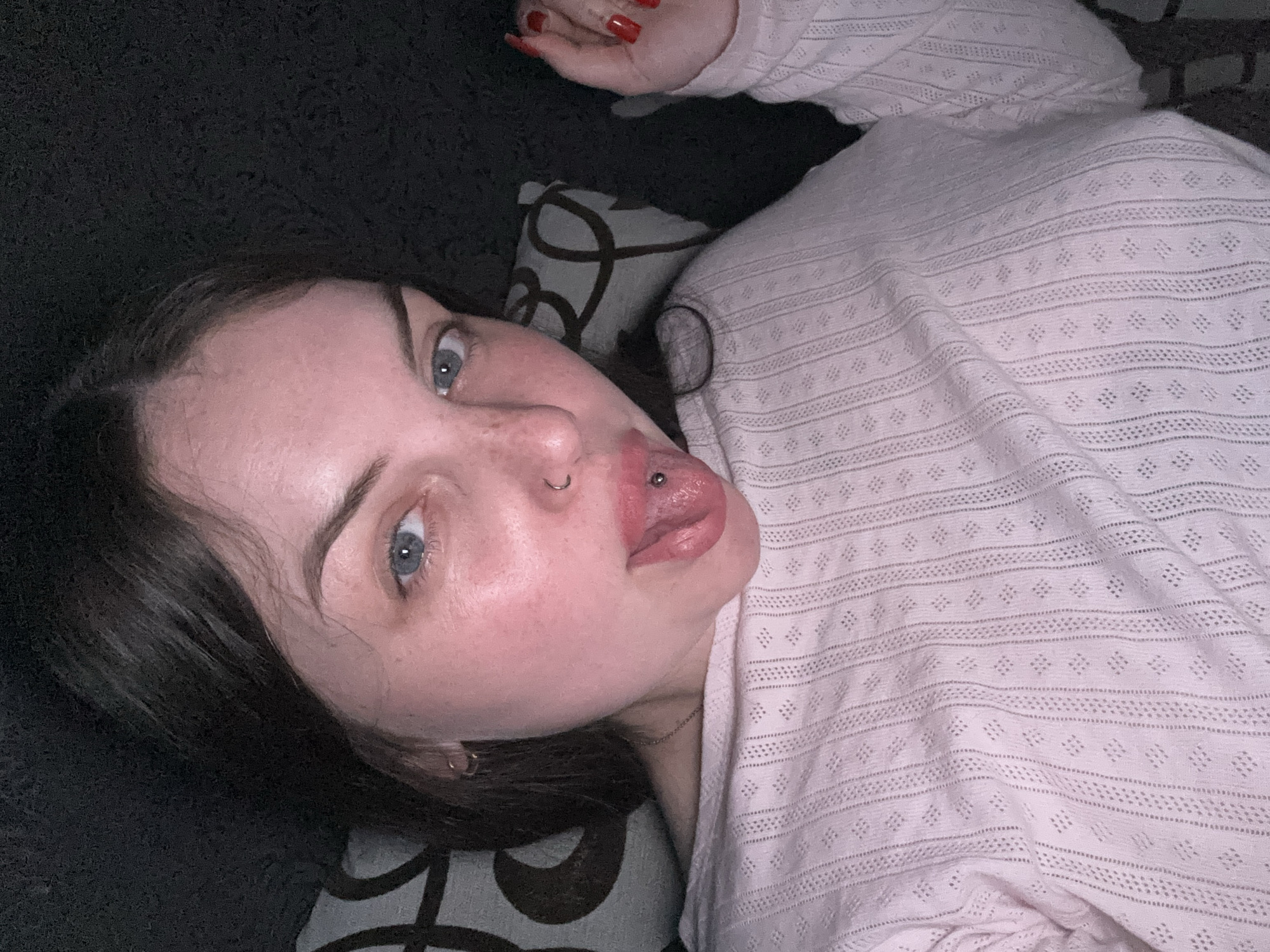 A young woman lies on a dark, patterned couch, her head resting on a pillow with a swirling design. She is wearing a pink, patterned long-sleeved shirt. She has fair skin, light blue eyes, dark hair, and a piercing in her tongue and nose. Her red fingernails are visible in the upper right corner of the frame. The background is a dark, textured material. The location and specific actions or interactions are not discernible from the image.