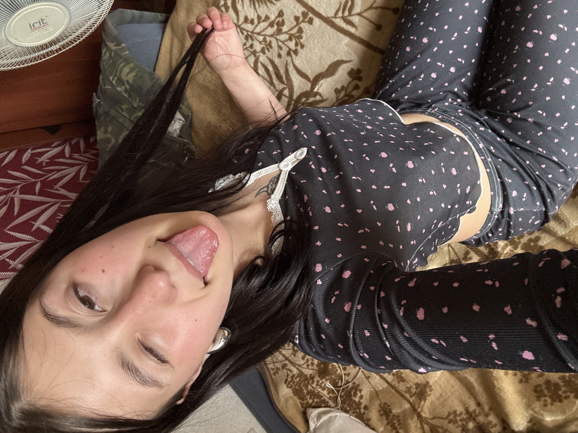 A young woman is depicted in a playful, close-up selfie, lying down in an indoor setting. She has long, dark hair, brown eyes, and a fair complexion with a few small freckles on her nose and cheeks. Her expression is lighthearted, as she winks her right eye and sticks out her tongue. She is wearing black pajamas with a scattered pink dot pattern, the top featuring white lace trim and a white decorative clasp on the chest. Her left hand is visible near the top of the frame, gently pulling a strand of her hair.
The background shows various textures and objects. To the upper left, a white electric fan with the brand name "IRIT" is clearly visible on its grille. Below the fan, a reddish fabric with a white leaf-like pattern and a dark green, camouflage-patterned fabric are visible. The woman is largely resting on a brown and gold patterned blanket or bedspread. A white earbud or wire is faintly visible near her midsection. The overall impression is one of casual comfort and fun, suggesting a relaxed moment in a home in Фоки, Russia. The presence of the fan might suggest a warm environment.