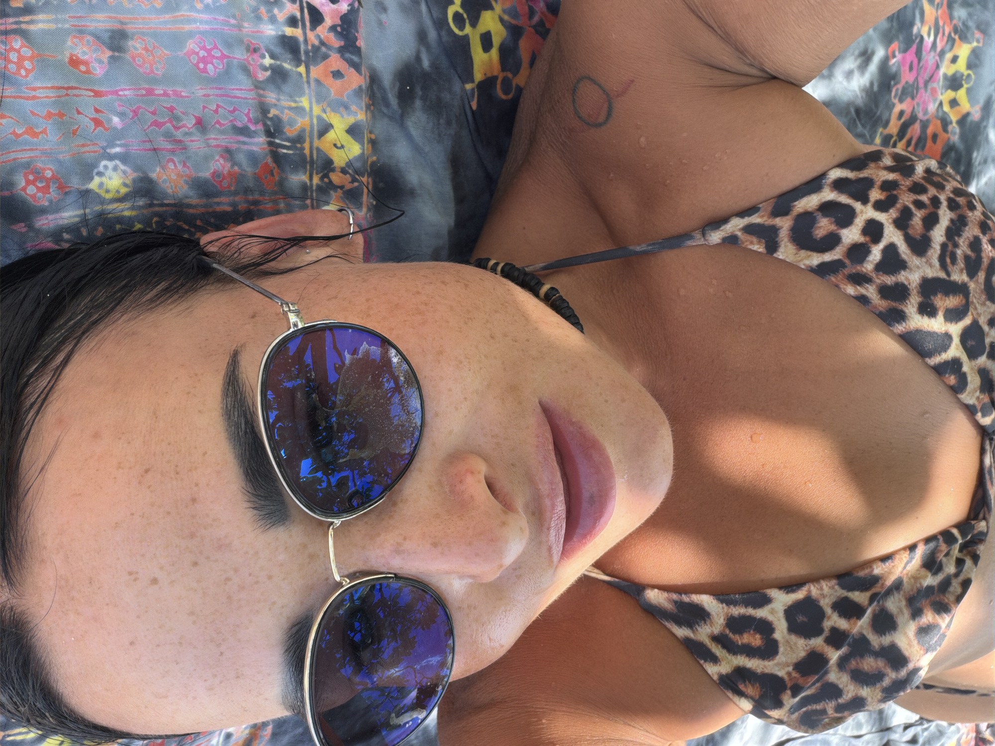A medium-close-up shot from a slightly elevated perspective shows a woman lying down on a patterned blanket. She is wearing round sunglasses that reflect trees in the lenses and a leopard-print bikini top. A small circular tattoo is visible on her neck. The location is likely Nusa Dua, Indonesia, given the general aesthetic and possible origin.