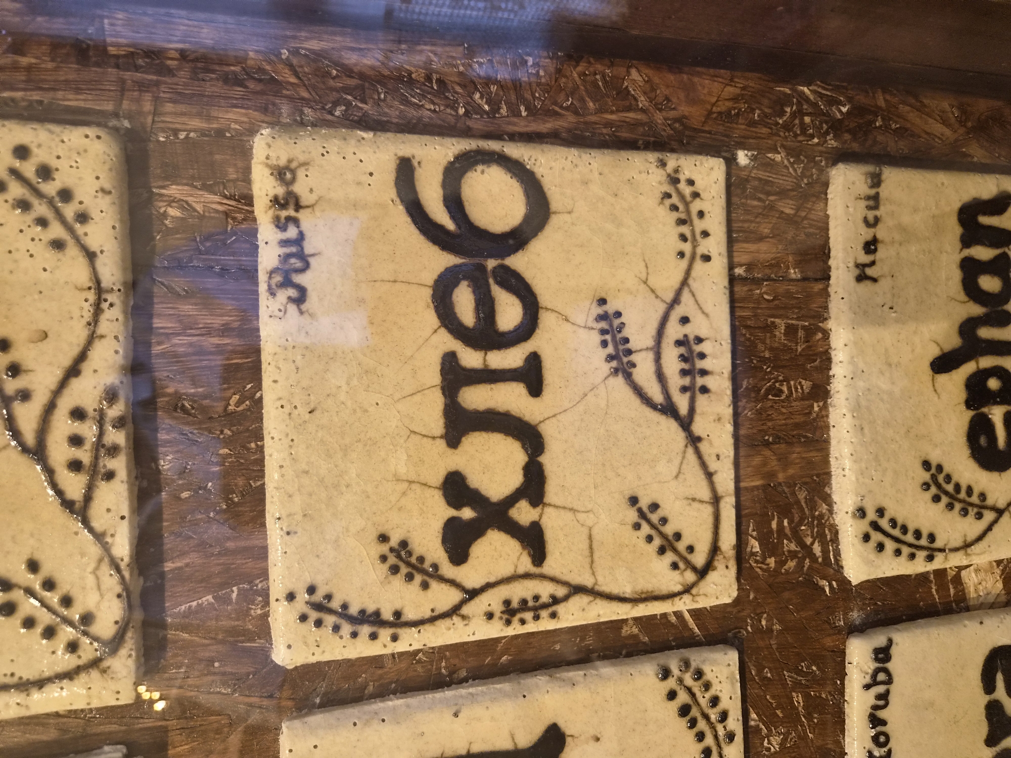 The image displays a close-up view of decorative tiles mounted on a wooden surface. The tiles are light-colored with a crackled glaze and feature dark, hand-painted designs. One prominent tile in the center has the Cyrillic word "хлеб" (khleb), meaning "bread," written in large, bold, black lettering. To the upper right of this tile, the word "Roussa" is written in a smaller, dark script. Another tile to the lower right shows the word "Hacua" in a similar script. Partially visible tiles on the left and bottom show partial words that are difficult to decipher clearly. The overall presentation suggests these are artisanal creations, possibly souvenirs or decorative items. There is no visible indication of the city of Seia, Portugal, within the image itself.