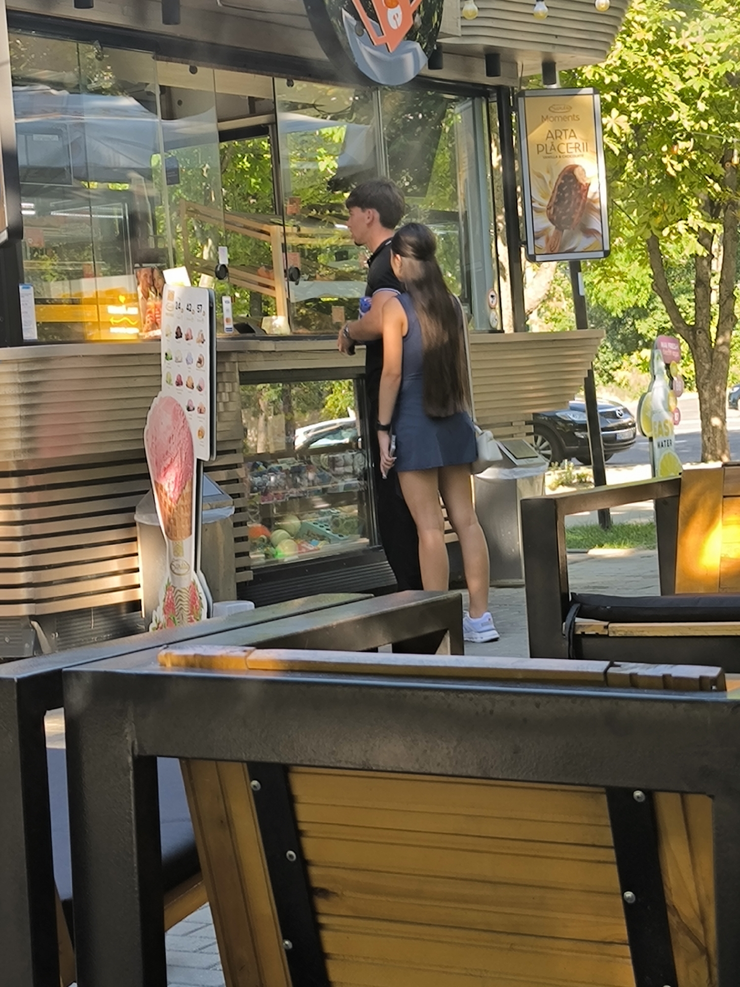 In Chișinău, Moldova, a couple stands near an ice cream kiosk. The woman, wearing a blue dress and white sneakers, has long dark hair. The man, in a black shirt, stands beside her. The kiosk displays various ice cream flavors and has a large advertisement for "ARTA PLACERII" (Art of Pleasure). Behind them, a black SUV is parked. The scene is set outdoors, with trees and sunlight contributing to a warm atmosphere.
