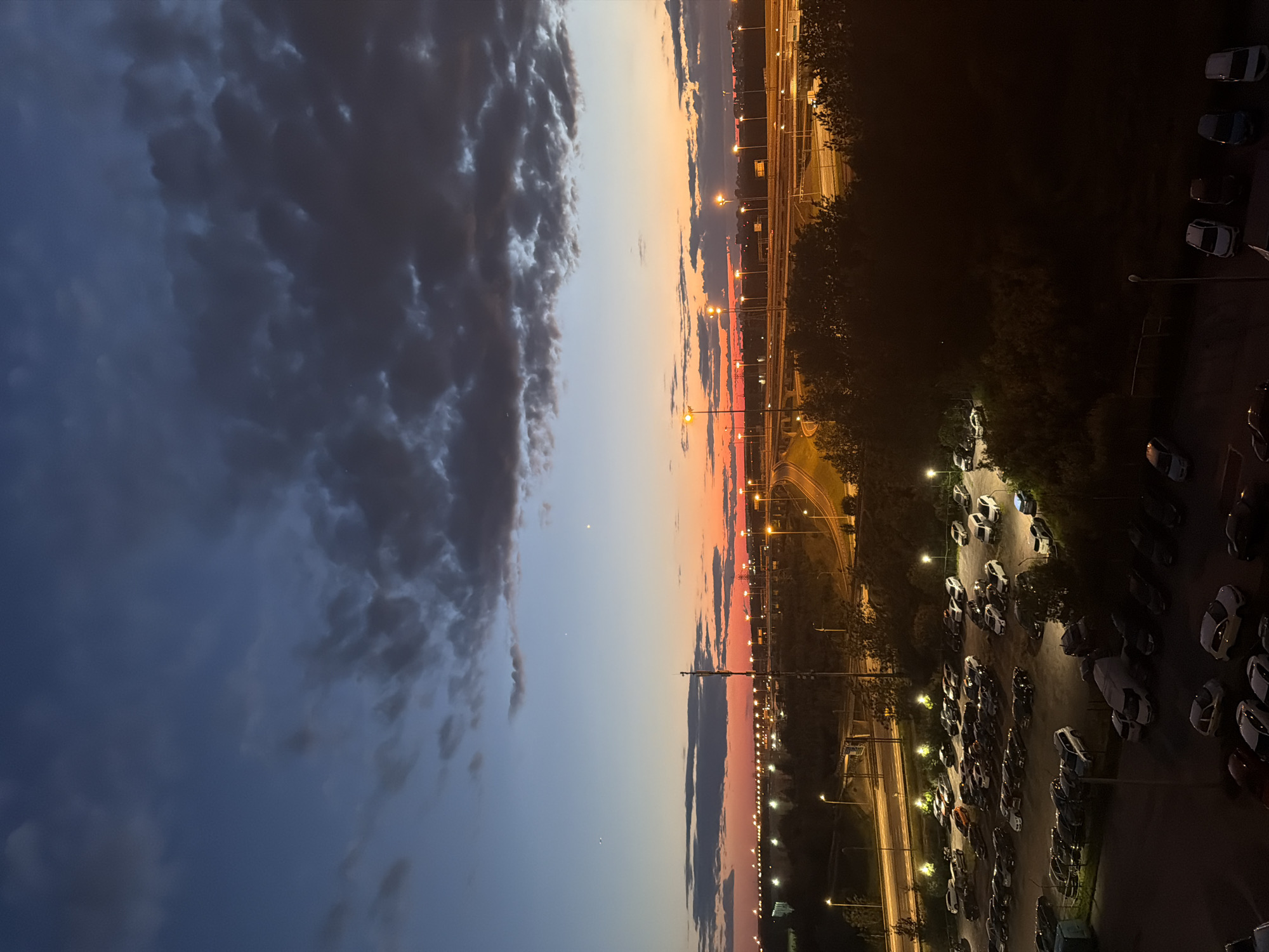 The image depicts a nighttime scene in an urban environment, possibly an airport or a large industrial area, given the presence of numerous parked vehicles and extensive road infrastructure. The sky is a gradient of deep blues and grays, transitioning to a vibrant orange and red sunset on the horizon, suggesting the time is either dusk or dawn. Dark clouds are scattered across the sky, illuminated by the ambient light.
In the foreground, a large, dimly lit parking lot is filled with a multitude of cars. The vehicles are various colors and models, parked in rows. Some of these cars are illuminated by nearby streetlights. To the left of the parking lot, a light blue shipping container is visible.
Behind the parking lot, a complex network of roads and overpasses dominates the midground. These roads are lined with streetlights that cast a warm glow, illuminating the asphalt and surrounding trees. The trees are dark and silhouetted against the brighter lights and sky, with dense foliage.
In the distance, the city skyline is visible, marked by a continuous line of bright lights, possibly from buildings or further industrial activity. The overall impression is one of a vast, functional urban landscape transitioning from day to night, or vice versa, with a focus on infrastructure and transportation. There are no people visible in the image.