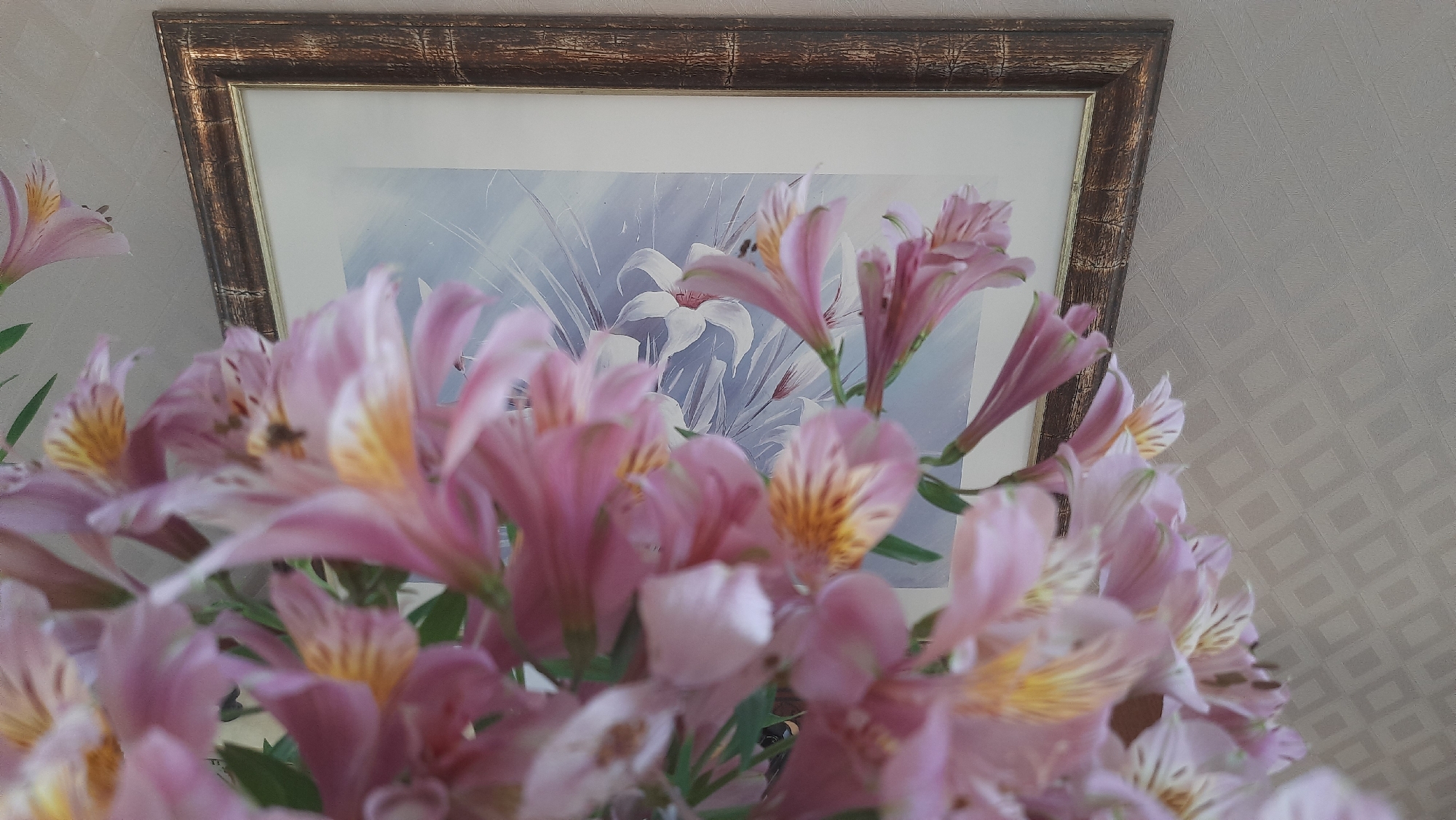A close-up, low-angle, indoor shot features a bouquet of pink alstroemeria flowers in the foreground, some of which are slightly out of focus. The flowers have yellow and brown markings on their petals. Behind the flowers, a framed picture is visible, depicting white lilies against a pale blue and white background with suggestions of tall grass. The frame is dark brown with a textured surface. The background wall to the right of the picture has a textured, diamond-patterned wallpaper in a light neutral color. The scene is likely a domestic setting in Desnogorsk, Russia, possibly a living room or dining area.