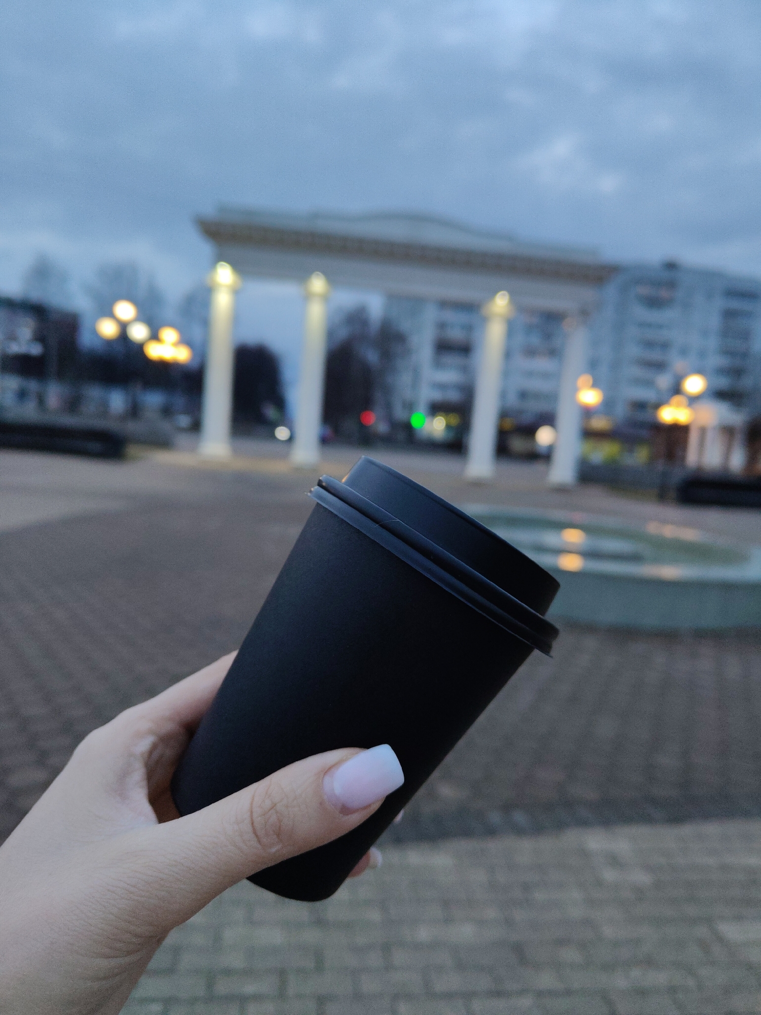 A photo shows a hand holding a black coffee cup in front of a city square. The hand, with pale skin and long white and pink fingernails, firmly grasps the cup. The cup, also black, has a lid and appears to be made of thick paper or cardboard.
In the background, slightly out of focus, is a city square made of light gray brick. A white archway structure with columns stands prominently in the center. Behind the archway is a multi-story building. Several round lamps on decorative poles light the square. A fountain is also visible, reflecting the lights in its surface. The sky is overcast, suggesting it may be evening or early morning. The location is Unknown, Unknown.