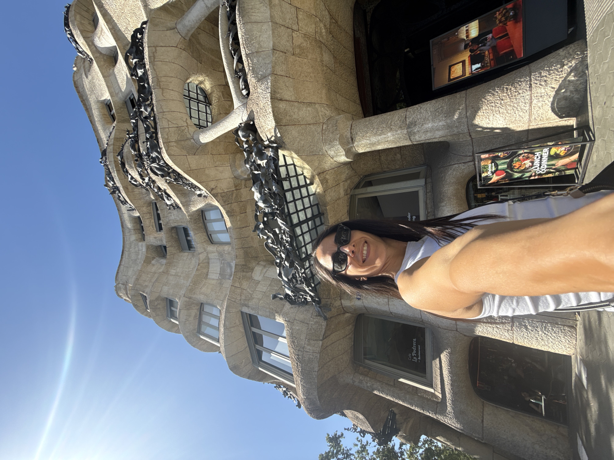 A medium-shot, wide-angle selfie captures a woman with long dark hair and sunglasses posing in front of a distinctive wavy stone building. The building, identified as Casa Milà, also known as La Pedrera, located in Barcelona, Spain, exhibits an organic, undulating facade with unique balconies featuring dark, sculptural elements. The woman is wearing a white tank top and is smiling at the camera, extending her right arm to take the photo. In the background, the building's architecture is prominent, with multiple floors and windows visible. Signs for "Café La Pedrera - Restaurant" and other promotional displays are situated near the entrance. The sky is clear and bright blue, indicating a sunny day. Trees are visible to the left of the building.