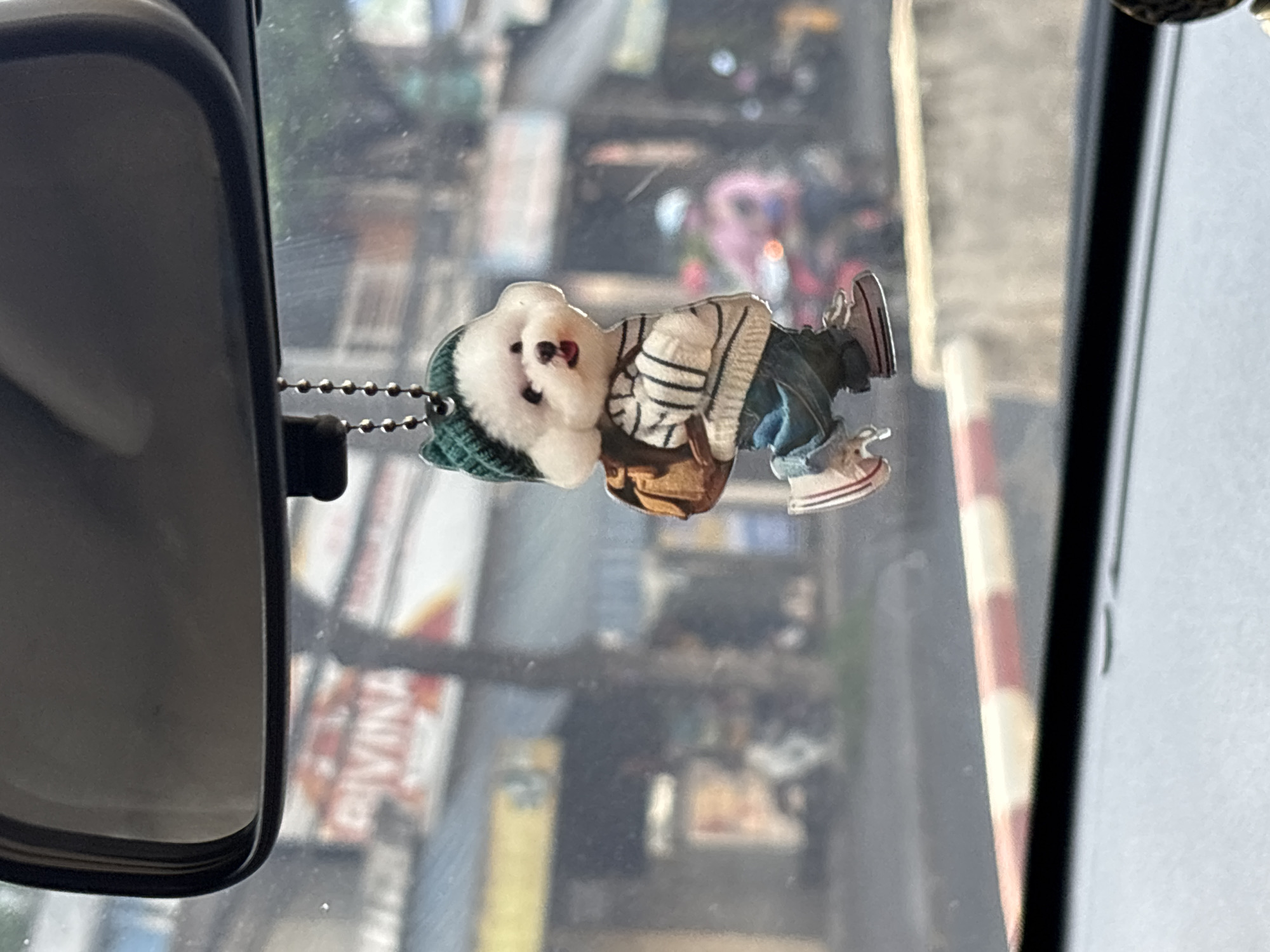 A small, fluffy white dog figurine wearing a green knit hat, a striped sweater, blue jeans, and sneakers is hanging from a beaded chain, suspended from the rearview mirror of a vehicle. The figurine has a brown backpack slung over its shoulder and appears to be mid-stride. In the background, seen through the car's windshield, is a street scene in Nha Trang, Vietnam. The street is lined with buildings, and there is traffic, including people on motorcycles. A red and white striped barrier is visible in the foreground. The overall impression is of a whimsical car accessory against a bustling urban backdrop.