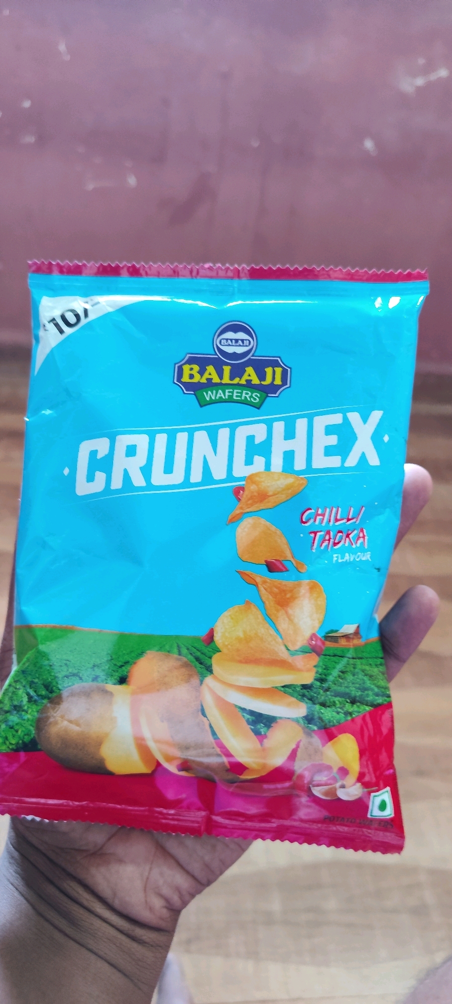 A person's hand is holding a blue and red bag of Balaji wafers, specifically "Crunchex" in "Chilli Tadka" flavor. The bag has an image of potatoes and chips. The background appears to be a wall with some subtle marks. The scene is likely indoors, given the lighting and the presence of a wooden surface below. No specific location in Belagavi, India is identifiable from the image.