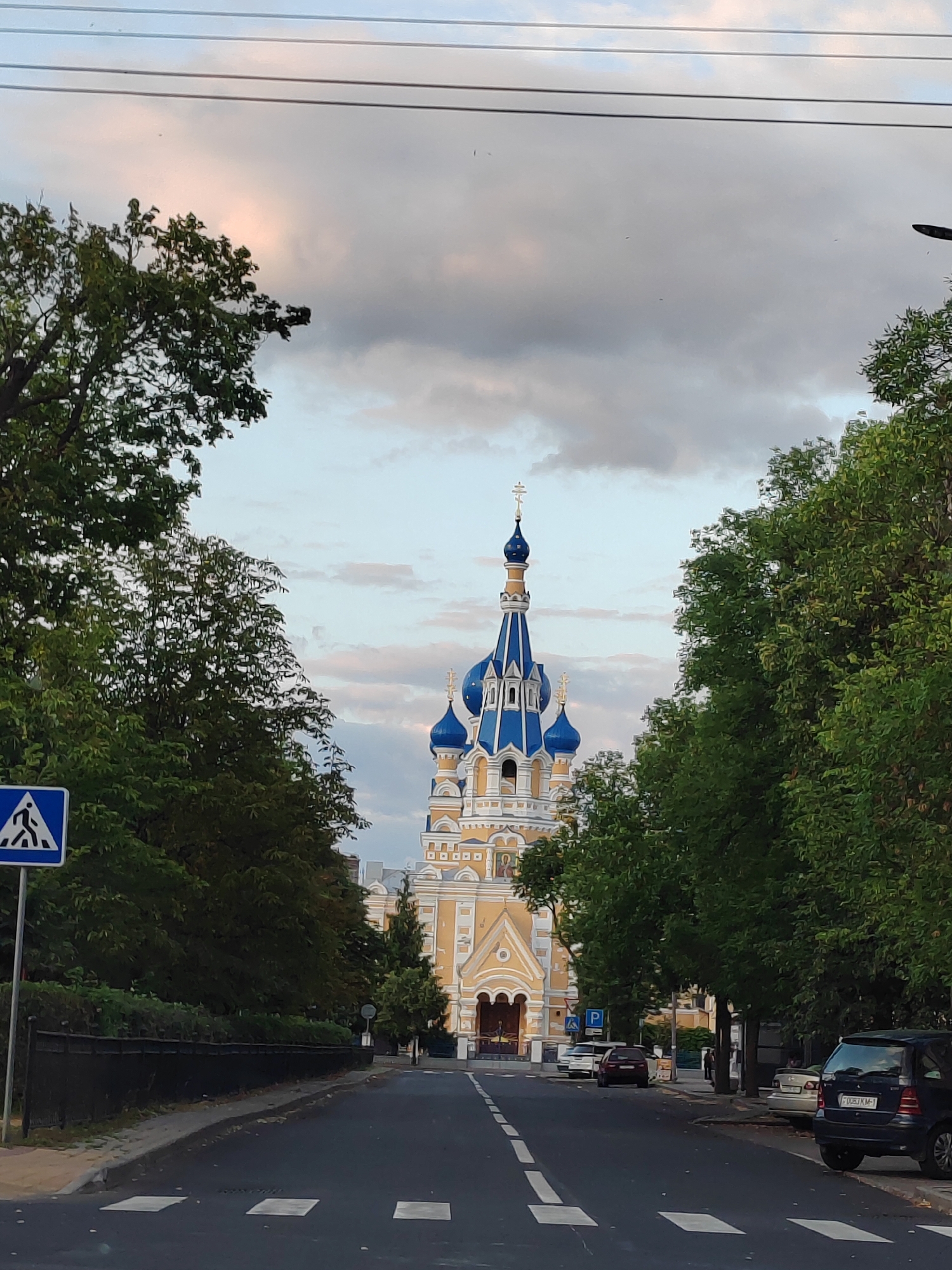In Brest, Belarus, a street scene captures an Orthodox church with blue domes and yellow facade, flanked by lush green trees. Overhead wires crisscross the sky, which features a mix of light and dark clouds, with hints of pink. A pedestrian crossing is visible on the asphalt road, marked by white stripes. To the left, a blue and white sign indicates a pedestrian crossing. Several cars are parked along the right side of the road. The overall impression is a quiet urban moment, with the majestic church as the central focal point.