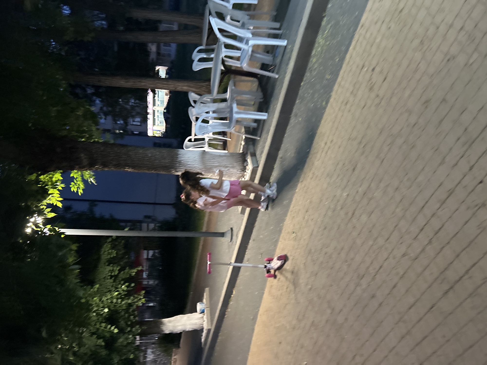 This image captures an outdoor urban scene at night in Dzhebel, Bulgaria, illuminated by artificial light. The foreground features a light-colored paved surface, possibly interlocking bricks or pavers, laid in a diagonal pattern. To the left, a children's three-wheeled scooter, predominantly white with pink accents on its wheels and handlebars, rests upright on this paved area.
Mid-ground, two young girls are standing near the base of a tree. The girl on the left wears a light pink top and shorts, while the girl on the right wears a white top and pink shorts and appears to be holding a small, light-colored object in her left hand. Both have long, dark hair.
A row of mature trees with dark trunks and foliage runs along the right side of the image. The base of at least two tree trunks is painted white. Beneath these trees, an outdoor seating area is visible, comprising multiple white plastic chairs arranged around at least two round tables with dark bases.
A tall street lamp with a bright light fixture is positioned towards the center-left, providing the primary illumination for the scene, casting a yellowish glow on the tree foliage and paved ground. In the background, dimly lit buildings with windows are visible through the trees, one displaying a bright, possibly illuminated, sign or interior light. A darker asphalt surface, likely a road or path, is present beyond the paved area and curb. A small white bucket or container is situated at the base of a tree on the left.