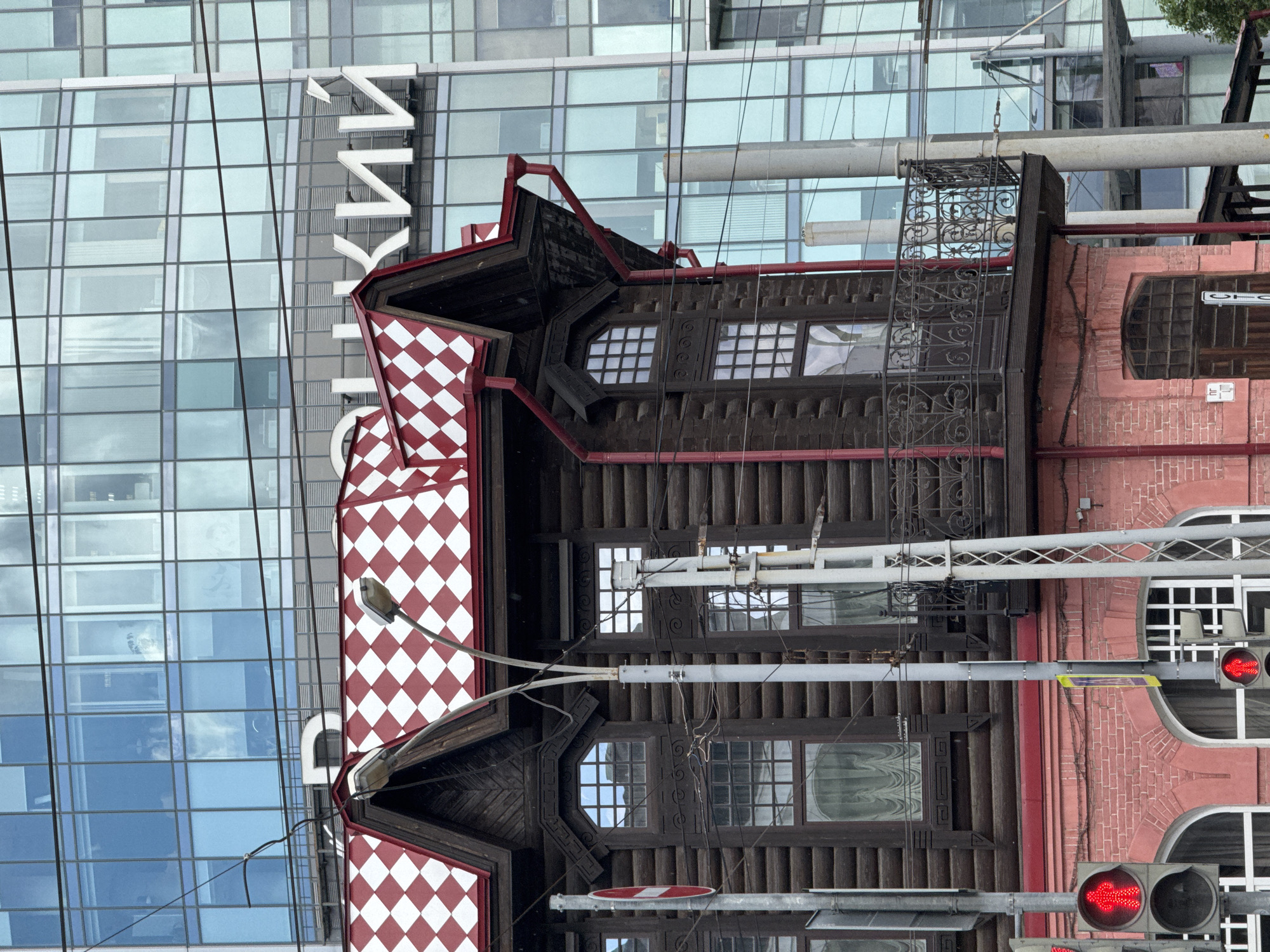 The image shows the exterior of a historic wooden building with a red and white checkered roof, juxtaposed against a modern glass skyscraper in Yekaterinburg, Russia. The wooden building features dark timber logs, ornate window frames, and a balcony with a decorative metal railing. Red gutters and downspouts are visible on the roof. The lower part of the wooden building is made of pink brick and has arched windows. A red traffic light showing a walking figure is visible on the left side of the frame. The glass skyscraper reflects the sky and surrounding environment, with visible office windows and some lettering. Utility poles and wires are also present in the foreground and midground. The scene captures a contrast between old and new architecture in an urban setting.