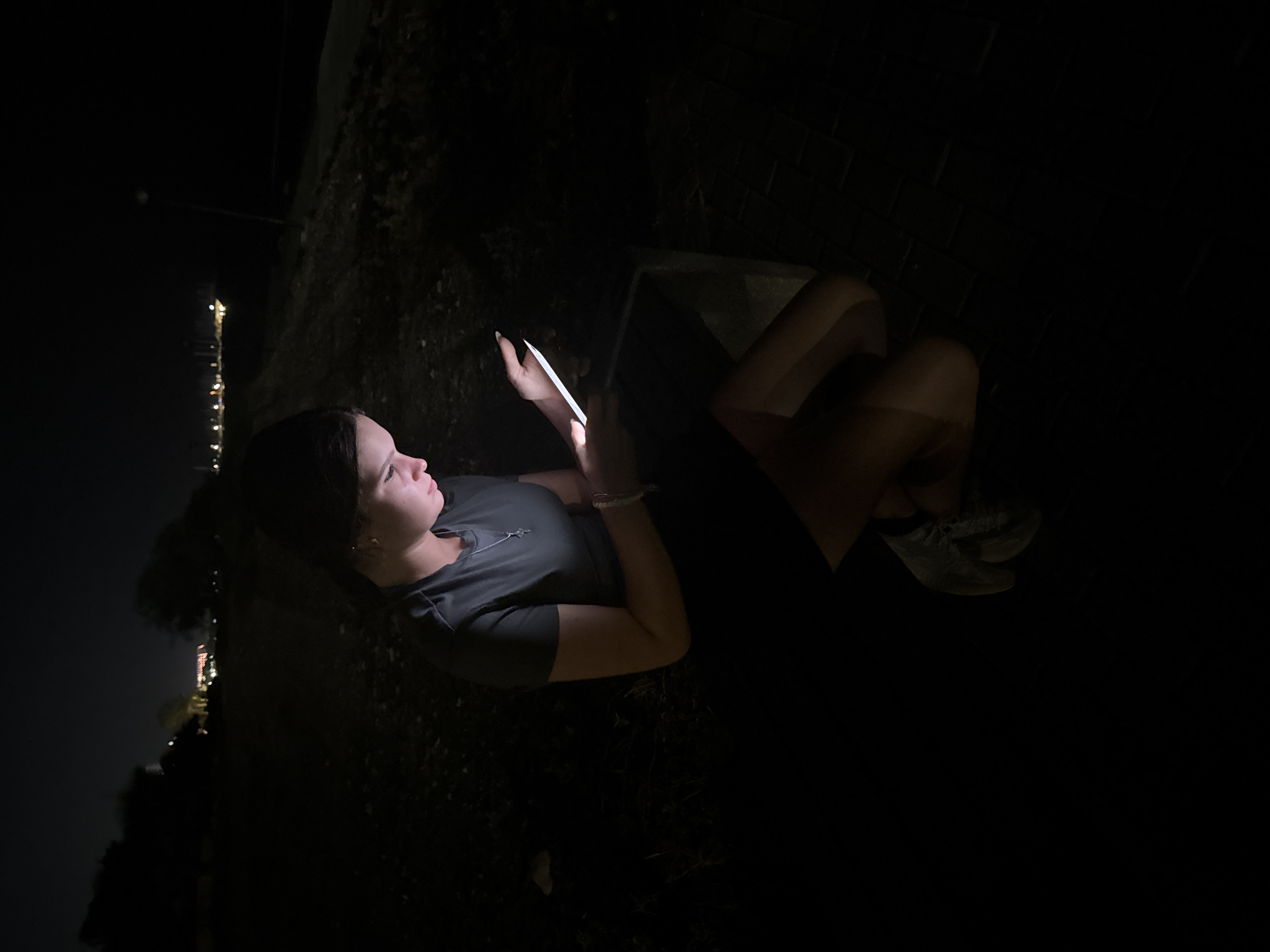 A young woman is crouched in the dark on a paved area next to a dirt path. She is illuminated by the light of a tablet device she is holding. She is wearing a dark t-shirt, shorts, and sneakers. In the background, distant lights from a town or city are visible under a dark sky. The scene appears to be outdoors, possibly in a park or along a walkway, near the coast of Parekklisia Community, Cyprus.
