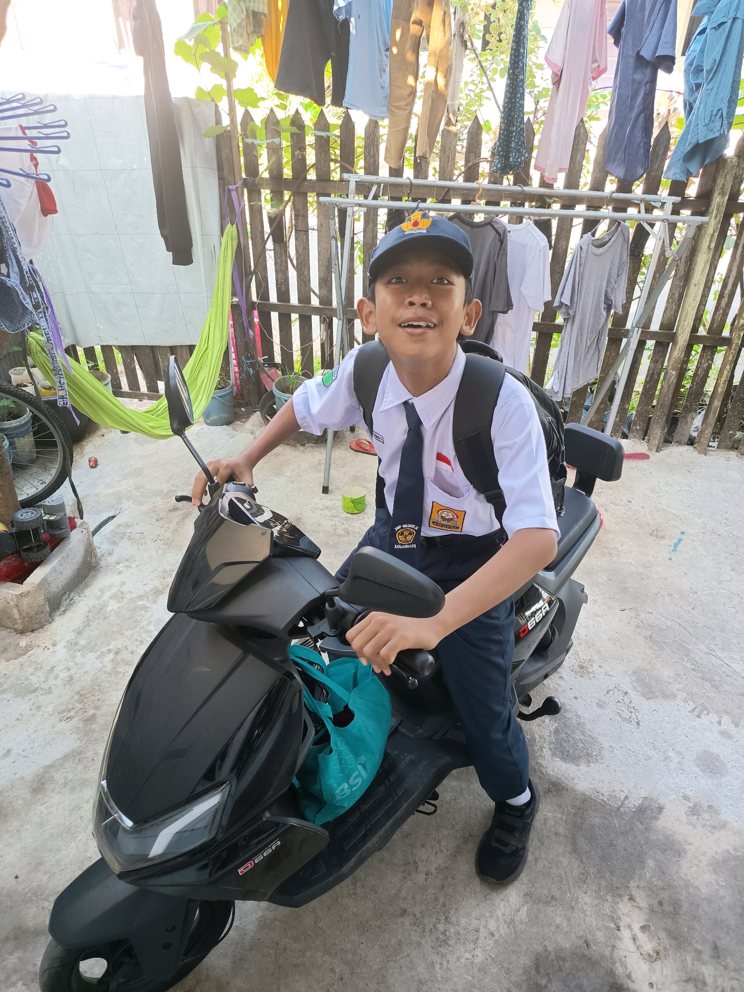 A young Indonesian boy, appearing to be of elementary school age, is seated on a black scooter. He is wearing a school uniform consisting of a white short-sleeved shirt, a dark tie with a school crest, navy blue pants, and a blue baseball cap. He has a black backpack on his shoulders and is holding the scooter's handlebars. A turquoise reusable shopping bag is hanging from the scooter's front. The background features a wooden fence with laundry hanging on clotheslines above it, indicating an outdoor residential setting. The ground is paved with concrete. A bicycle with a green hammock attached is visible on the left. The lighting suggests it is daytime. The school crest on the boy's uniform appears to be from "SMAN 2 Banjarmasin," placing the scene in Banjarmasin, Indonesia.