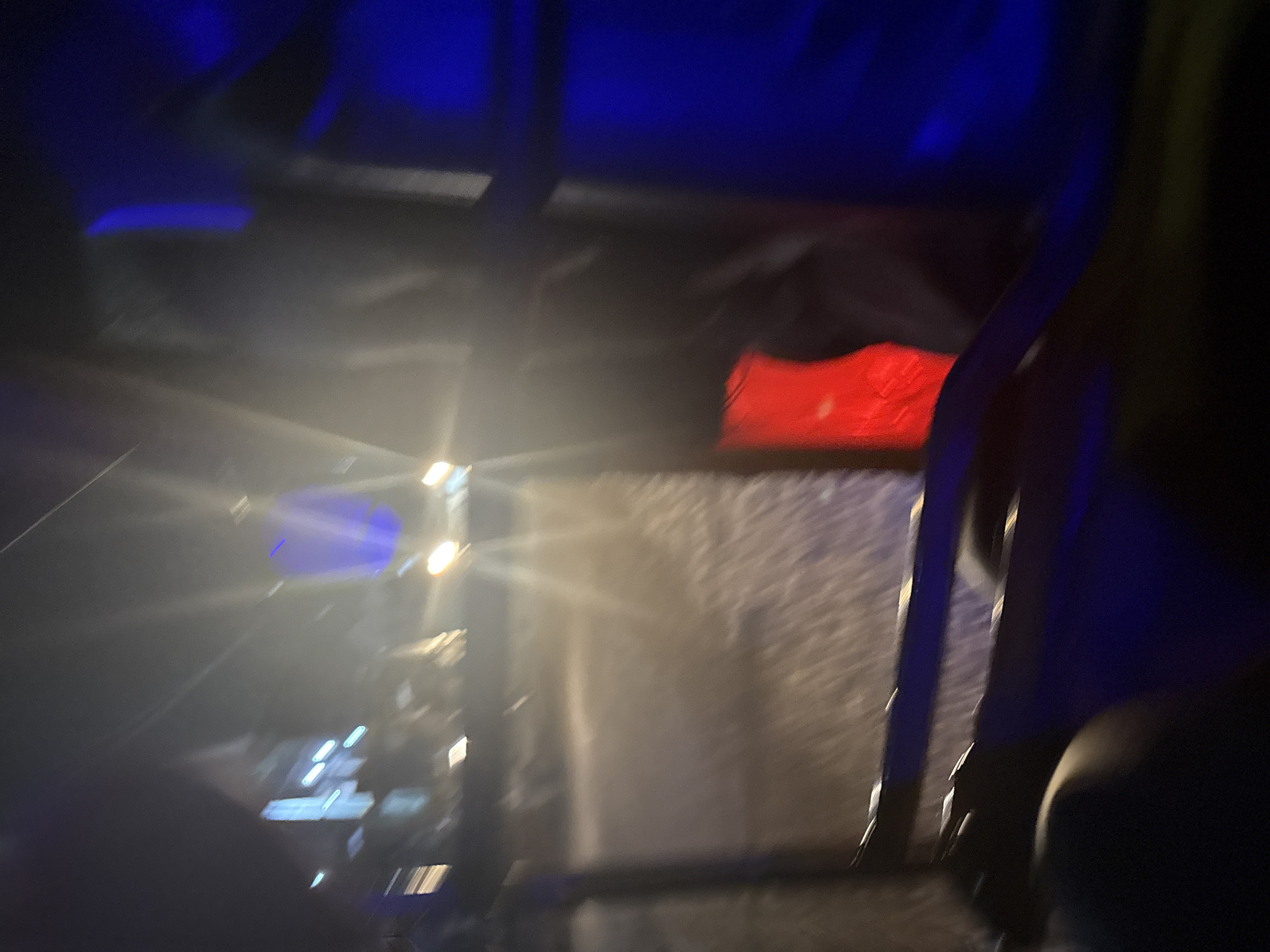 The image captures a blurry, nighttime scene possibly from within a vehicle. A prominent red taillight is visible on the right side of the frame, suggesting the presence of a vehicle. Blue lighting emanates from the left and right sides, casting an ethereal glow on the surroundings. Streaks of light suggest motion, possibly from streetlights or other moving vehicles. In the background, indistinct shapes of buildings and lights indicate an urban or semi-urban environment, consistent with Nkpor, Nigeria. The overall impression is one of a transient, dimly lit moment in a bustling city.