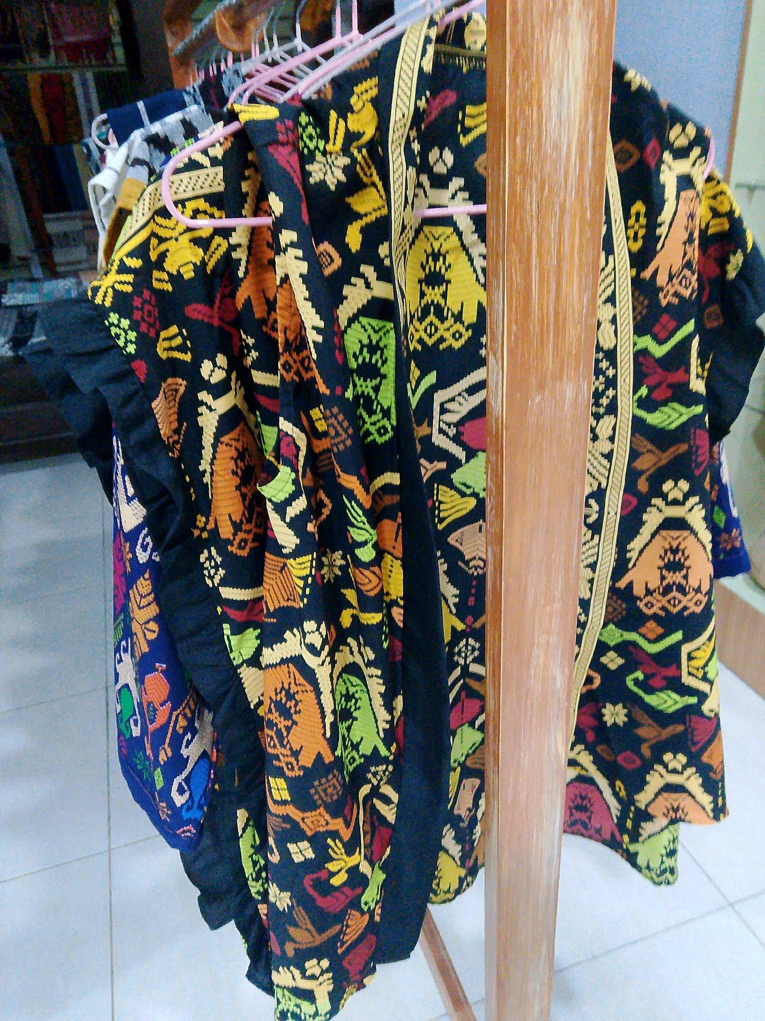 A close-up, eye-level shot shows several pieces of vibrantly patterned, woven fabric hanging on pink plastic hangers from a wooden display stand. The fabrics are predominantly black with intricate, geometric patterns in shades of yellow, orange, green, red, and blue. One piece, a vest or jacket, is adorned with what appear to be stylized animal figures and decorative motifs. The fabric is draped and folded, creating a sense of depth and texture. The background is slightly blurred, but suggests an indoor setting with tiled flooring and potentially other merchandise on shelves in the distance. The lighting is even, highlighting the detail of the textiles.