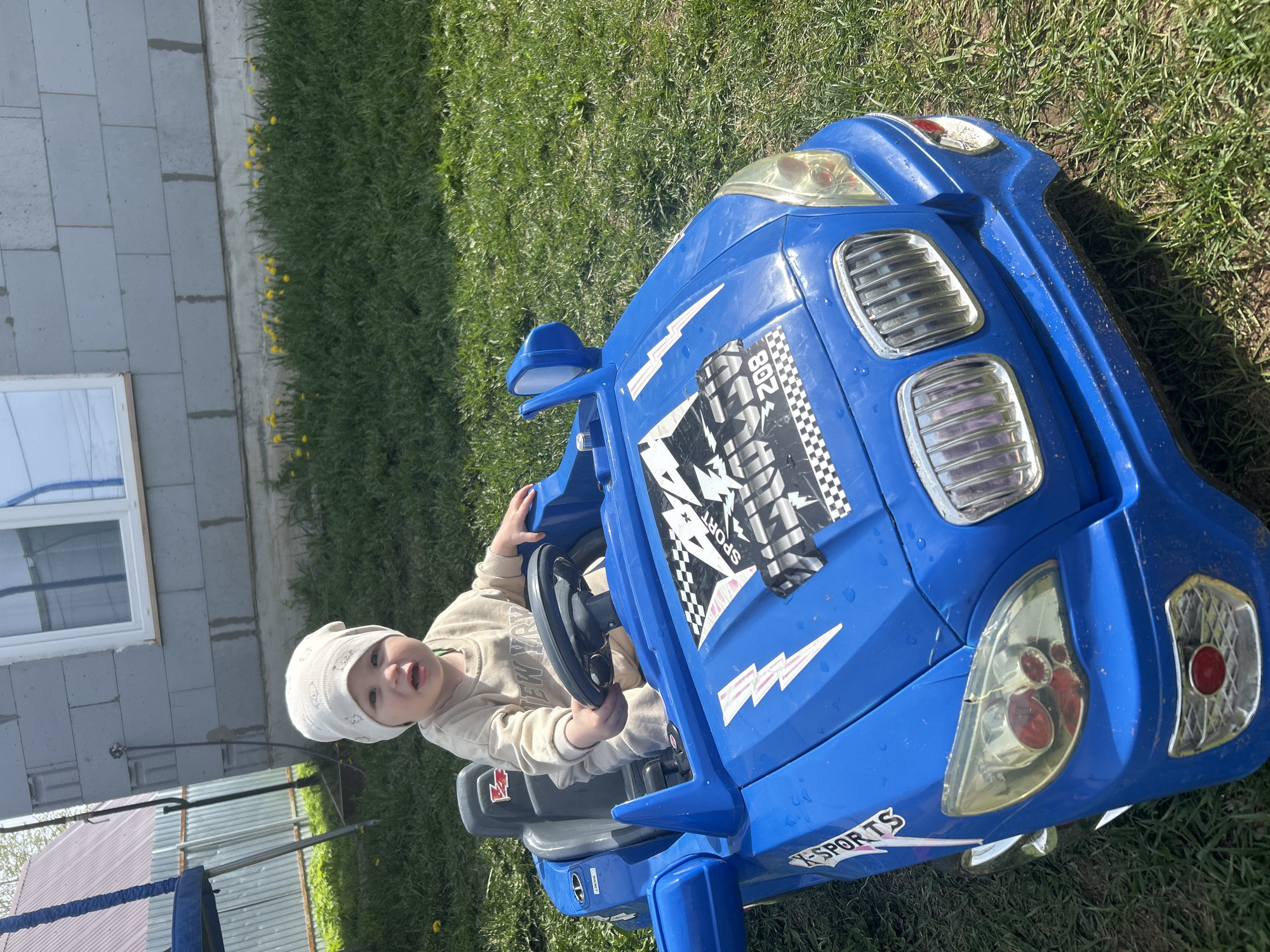 The image shows a young child seated in a blue toy car on a lawn. The child wears a light-colored hat and clothing. The car has stickers and design elements. The background includes a building with a window, grass, and what appears to be part of a trampoline frame. The image was taken during daylight. The city of Chaikovsky, Russia is not discernible from the image.