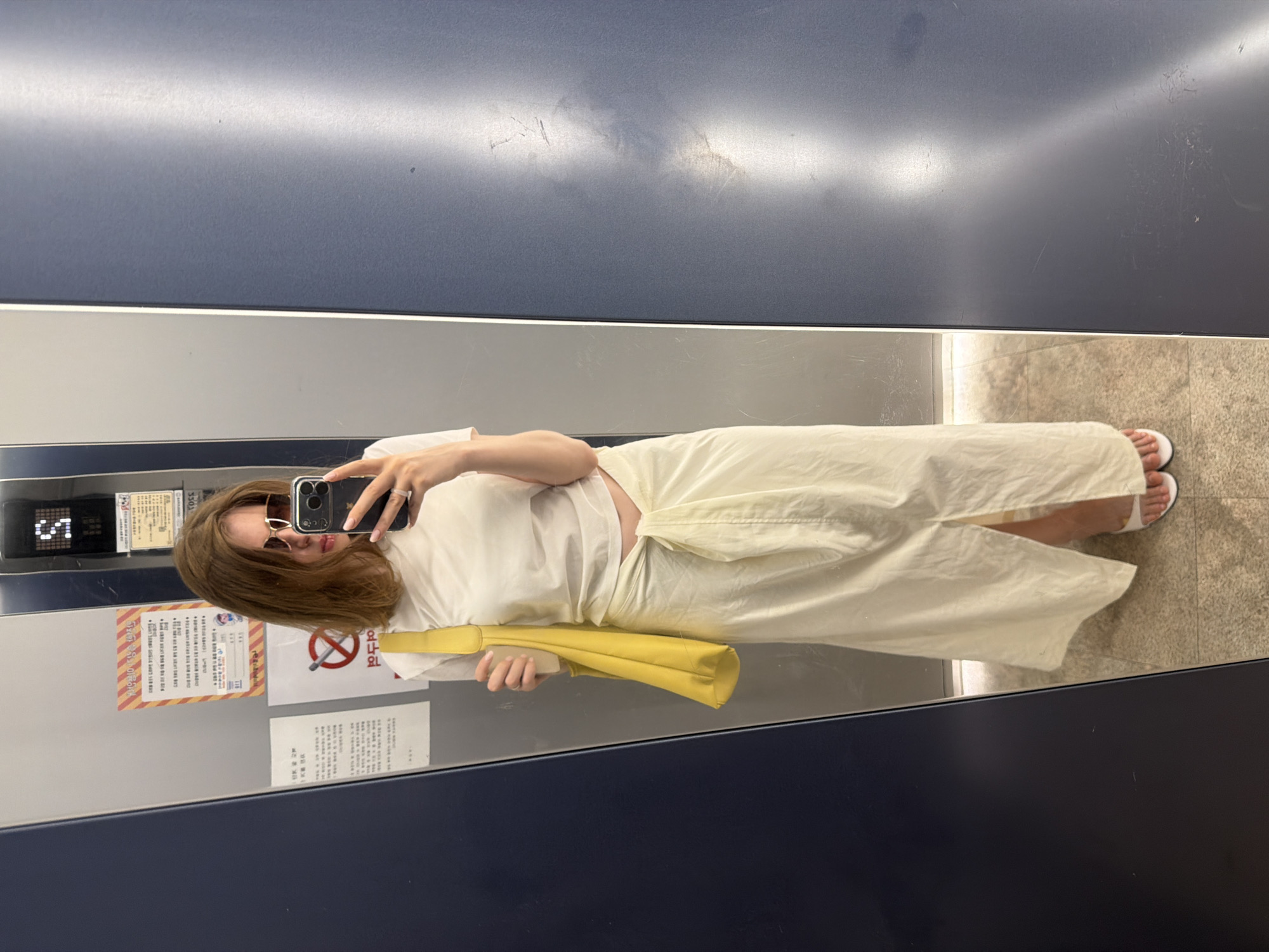 A woman with shoulder-length brown hair, wearing sunglasses, stands in an elevator, taking a selfie. She is wearing a white short-sleeved t-shirt and a long, cream-colored skirt with a tie detail at the waist and a slit up the front. She carries a bright yellow tote bag over her left shoulder. Her feet are clad in white slide sandals. The elevator walls are reflective, showing signs in Korean and a digital display with the letter "S" and a number. The floor is tiled. The location is the city of Dongducheon-si, South Korea.