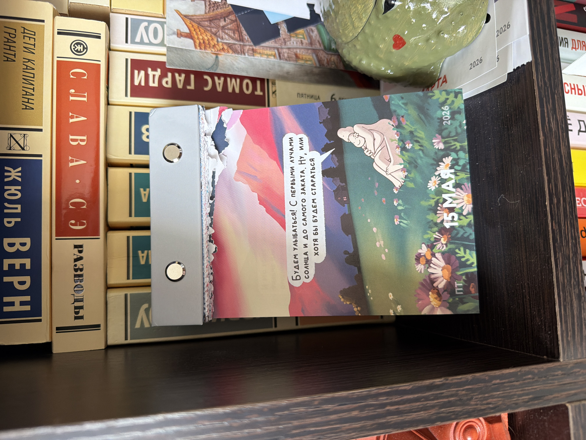 The image shows a shelf filled with books, primarily written in Russian. Prominently displayed is a tear-off calendar with an illustration and the date "15 MAY 2026" written on it. The calendar also contains Russian text, part of which translates to: "We will smile! With the first rays of the sun until sunset. Well, or at least we'll try". The books on the shelf appear to be older editions, with authors like Jules Verne ("Жюль Верн") and Thomas Hardy ("ТОМАС ГАРДИ") visible. There's a green ceramic object resembling a toad or frog positioned on top of some books. The scene suggests a personal library or bookshelf. The location is not discernable from the image.