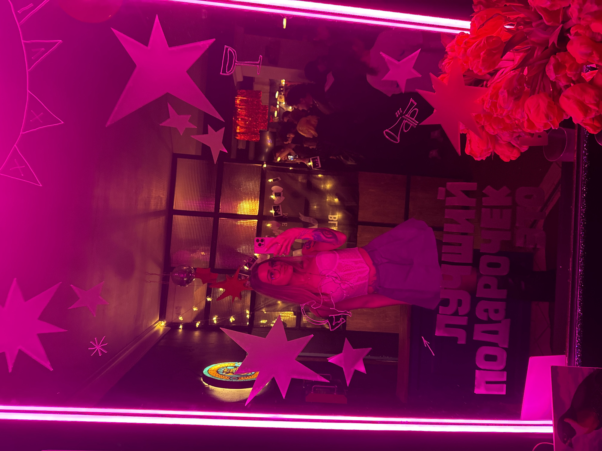 The image displays a mirror selfie taken by a young woman within an indoor entertainment venue, identified as being in Moscow, Russia, by the visible Russian text "ЛУЧШИЙ ПОДАРОЧЕК" (BEST GIFT).
**People:** A single female subject is prominent, positioned centrally in the reflection. She has long, straight, light brown hair, wears glasses, a white strapless corset-style top, and a light blue pleated mini-skirt. Tattoos are visible on her left arm. She holds a light pink smartphone, presumably an iPhone, to take the photograph. In the background reflection, a blurred crowd of multiple individuals is visible, suggesting a social gathering.
**Subjects and Objects:** The primary subject is the large mirror itself, which is decorated with numerous star-shaped cutouts, predominately in pink/magenta hues. Two vertical magenta neon light strips frame the left side of the mirror. Other neon-style outlines on the mirror depict a martini glass and a trumpet. The Russian text "ЛУЧШИЙ ПОДАРОЧЕК" is prominently displayed on the lower half of the mirror. In the lower right foreground, partially obscuring the mirror's surface, is a large bouquet of red-orange tulips.
**Environment and Lighting:** The setting is characterized by strong, pervasive magenta/pink neon lighting that dominates the scene and reflects off surfaces. Warm-toned fairy lights are strung horizontally in the background. Additional decor includes a small disco ball hanging from the ceiling, a faint banner with triangular pennants, a circular stained-glass-like fixture on the left wall, and a small white unicorn-shaped neon sign to the right of the woman's head. A section of windows or glass blocks is visible in the upper left background, and a red fringed decoration hangs from the ceiling further back. The overall environment appears to be an active, artificially lit indoor social space.