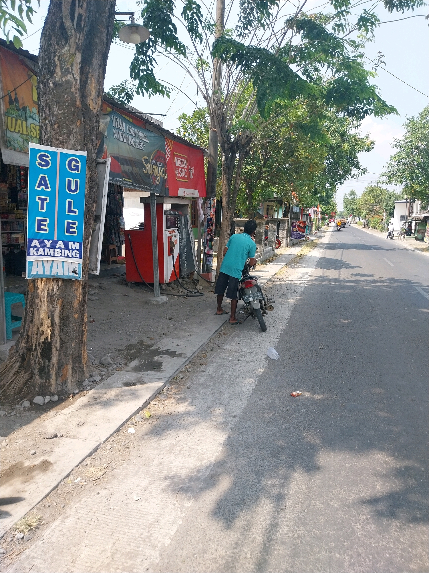 A man wearing a blue t-shirt and dark shorts stands on the sidewalk next to a motorcycle on a sunny day in Campur, Indonesia. The man appears to be interacting with the motorcycle. To his left, a tree trunk is visible with a blue sign that reads "SATE GULE AYAM KAMBING". Behind the man, there is a small roadside shop with a red gas pump. The shop has various items displayed and a banner that reads "TOKO ASTI SRC". The street is paved and runs alongside the sidewalk. Further down the street, there are other buildings and trees, and traffic can be seen in the distance. The environment appears to be a typical urban or semi-urban setting in Indonesia.